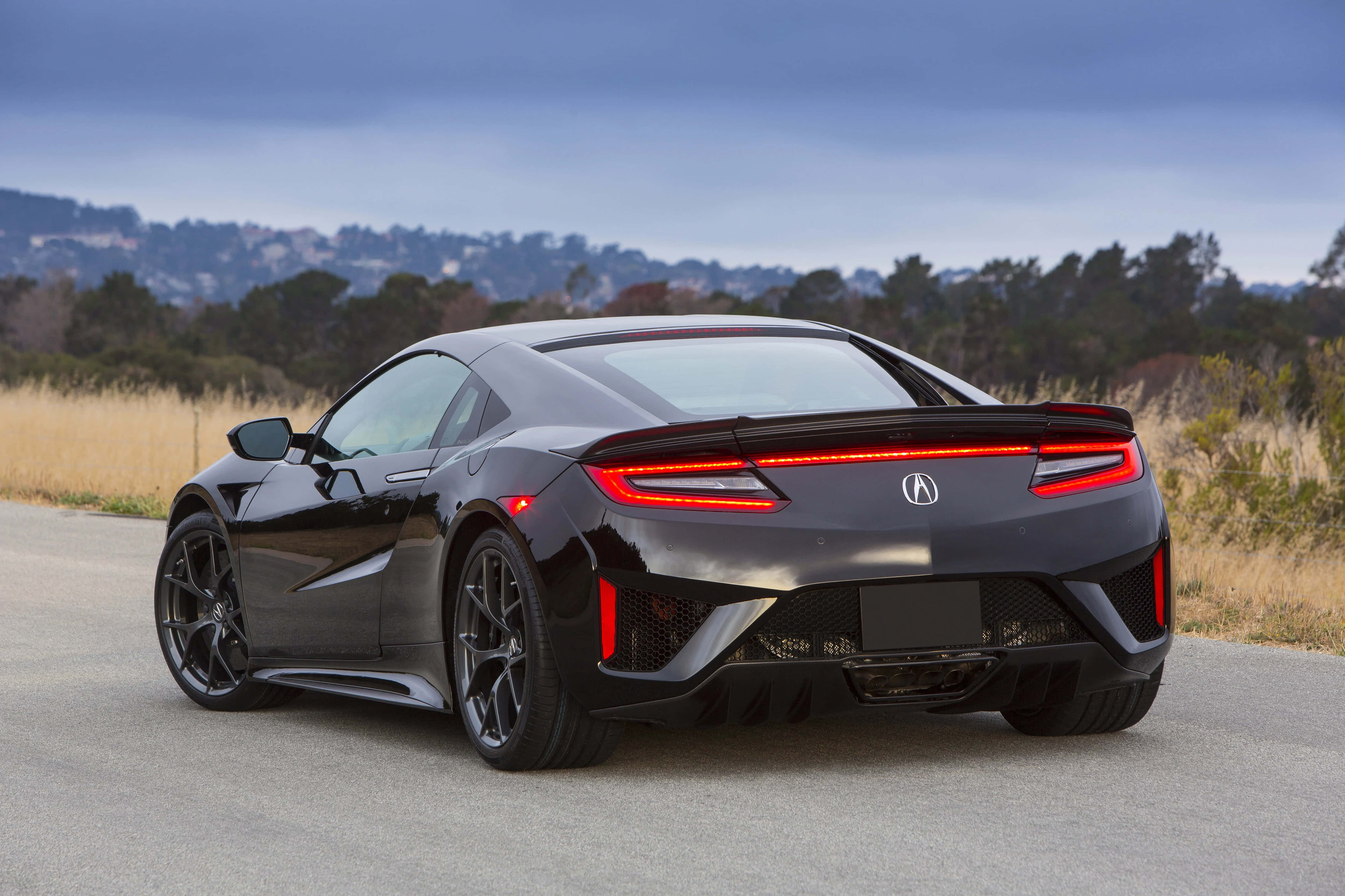 Honda Nsx 桌布 壁紙 — 一輛時尚的黑色跑車，帶有發光的紅色尾燈，停在一條有乾草和遠處山丘的道路上，天空多雲。