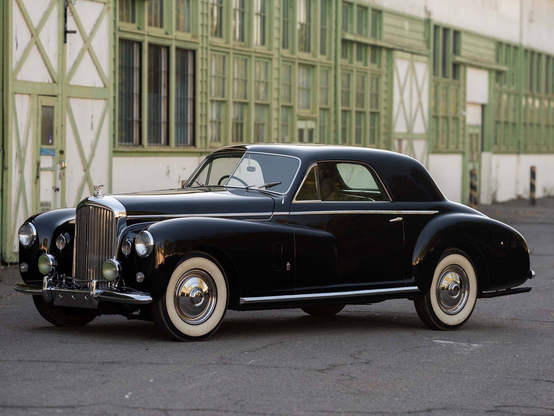 Bentley Mark Vi 桌布 壁紙 — 一輛時尚的黑色復古轎跑車，配備白色牆壁輪胎，停放在綠色和白色的工業建築前。
