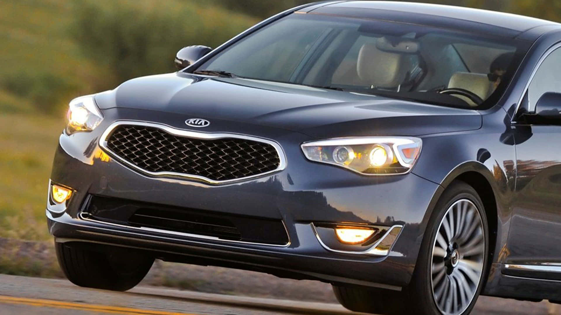 Kia Cadenza 桌布 壁紙 — 一輛時尚的深藍色Kia轎車，車頭燈閃爍，在陽光普照的道路上行駛，背景是模糊的綠植。