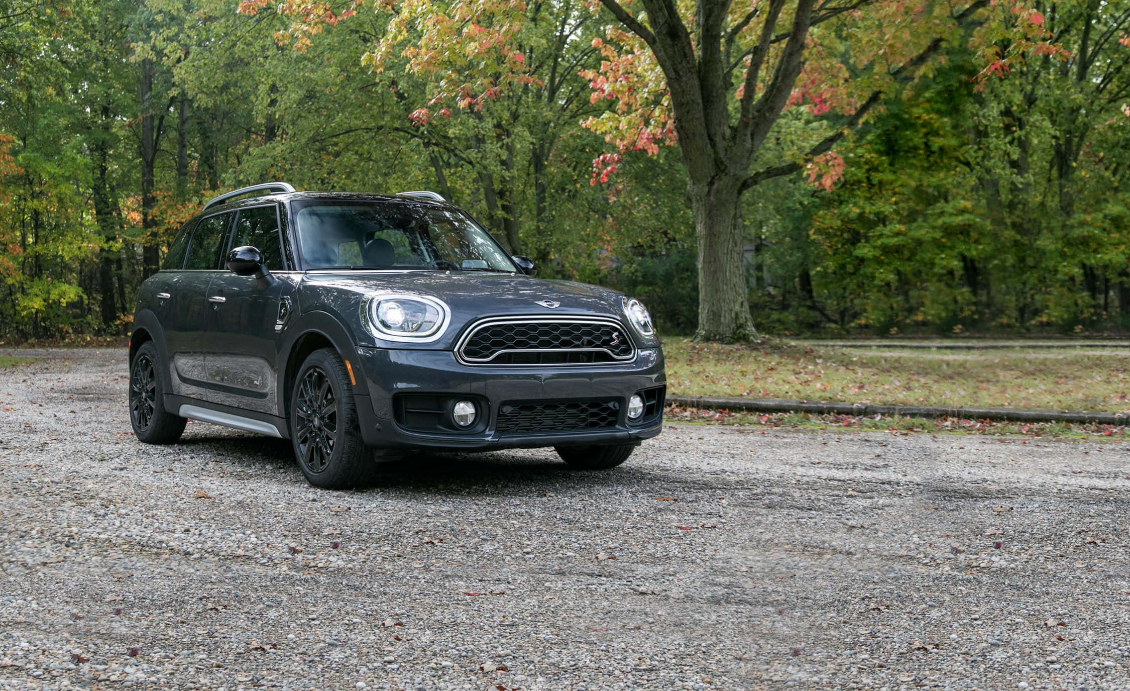 Mini Cooper S Clubman All4 桌布 壁紙 — 一輛時尚的深灰色MINI Countryman停泊在碎石路上，周圍是帶有橙色和綠色葉子的秋樹。