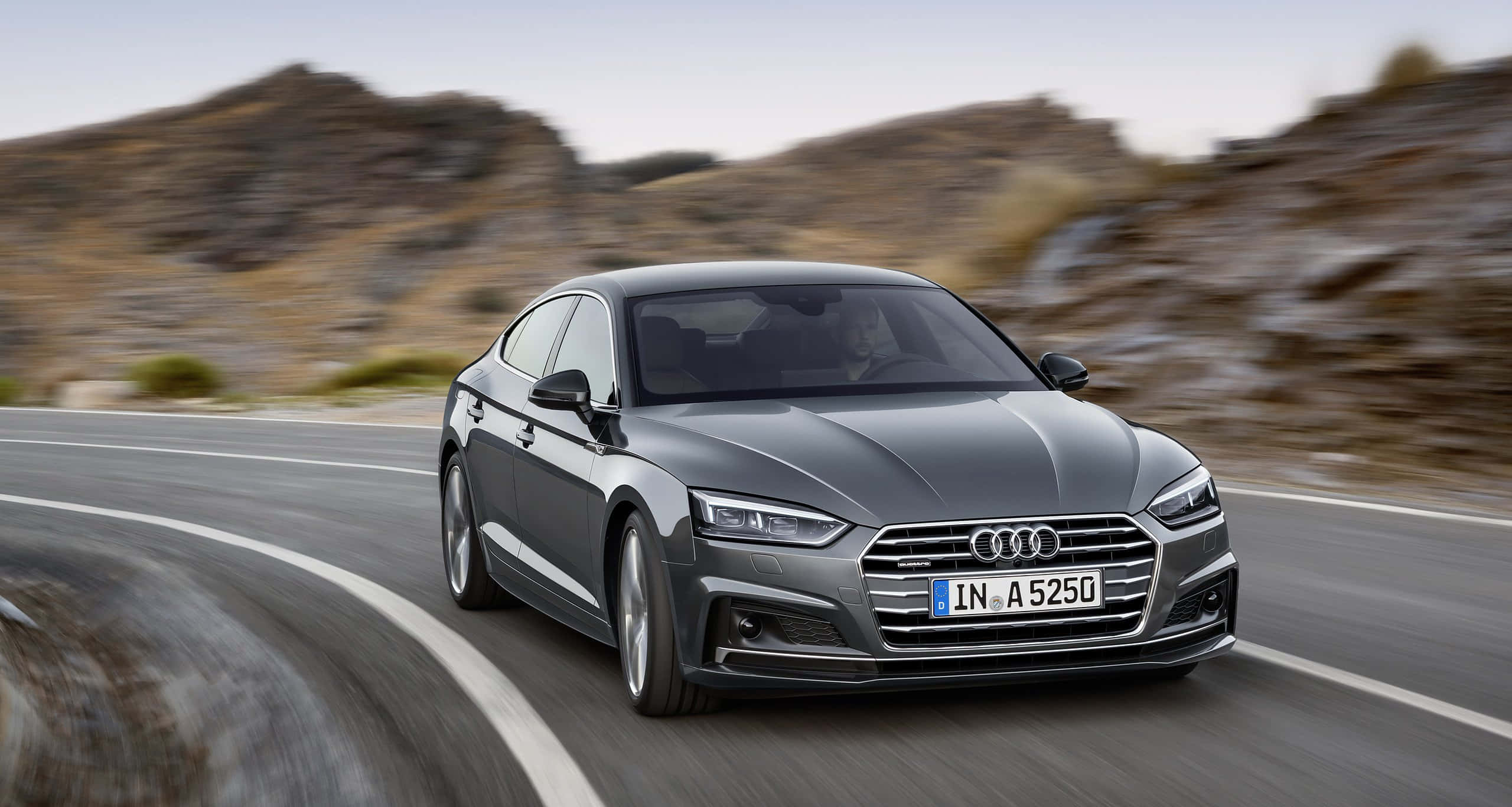 Audi A5 桌布 壁紙 — 一輛時尚的灰色奧迪轎車沿著蜿蜒的山路疾馳，背景是模糊的岩石地形。
