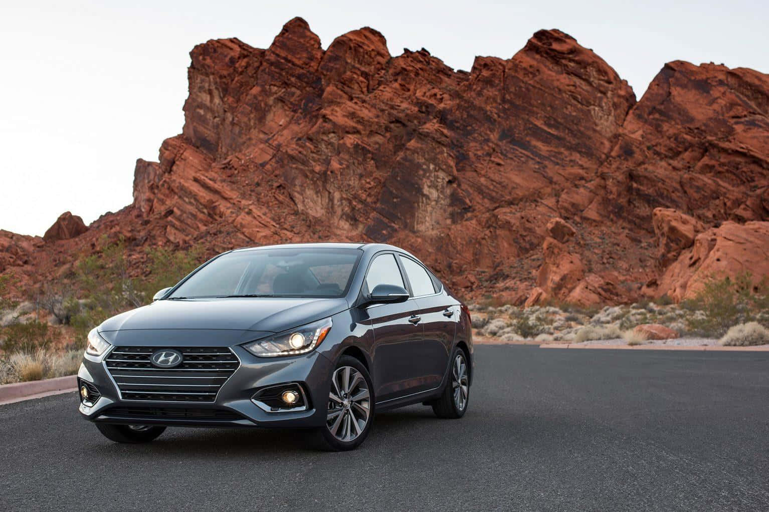 Hyundai Accent 桌布 壁紙 — 一輛時尚的灰色現代房車停在柏油路上，背後在蒼白的天空下聳立著戲劇性的紅色岩層。