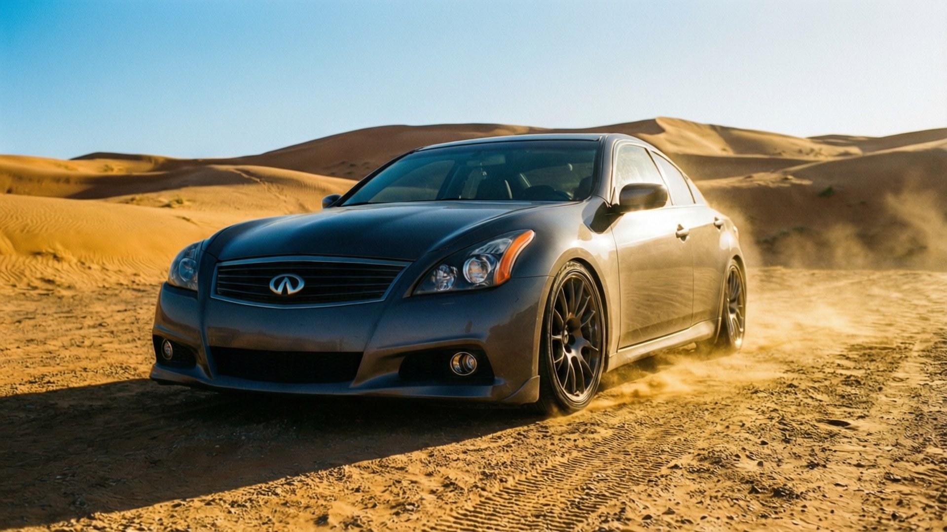 Infiniti G37 桌布 壁紙 — 一輛時尚的灰色 Infiniti 轎車在清澈藍天下，於金色沙漠沙丘上疾馳，揚起沙塵。