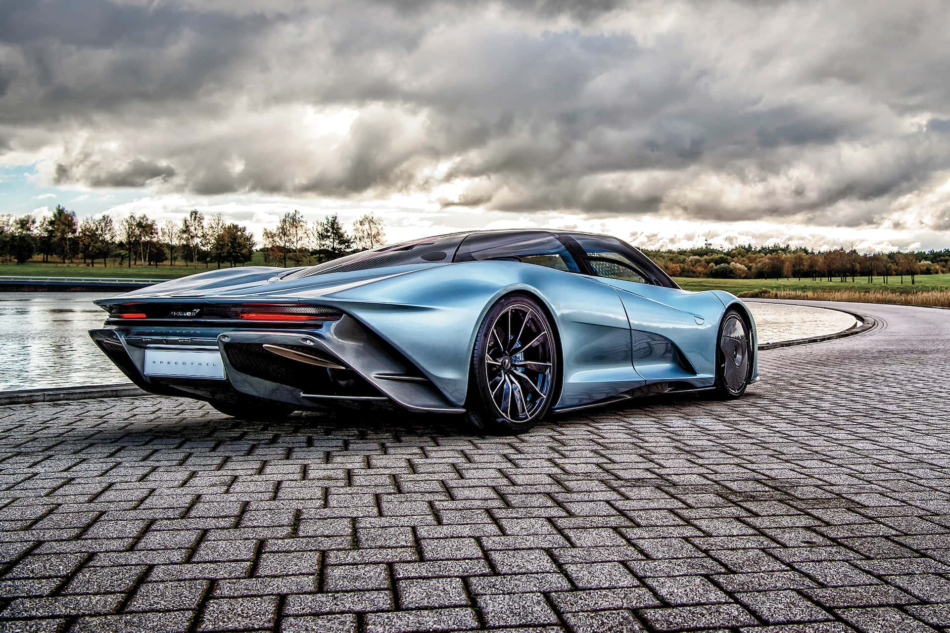 Mclaren Speedtail 桌布 壁紙 — 一輛時尚的淺藍色跑車帶有黑色點綴，停泊在平靜水道旁的鵝卵石小徑上，天空多雲而富有戲劇性。