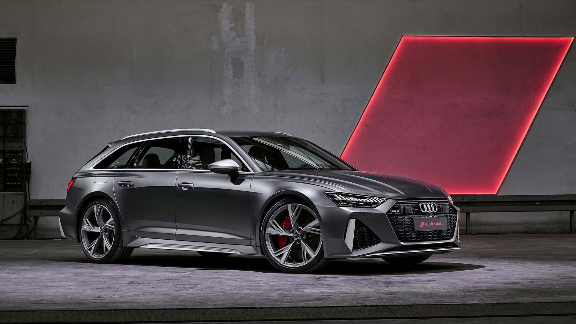 Audi Rs6 桌布 壁紙 — 一輛時尚的消光灰色奧迪RS6 Avant旅行車配備紅色制動卡鉗，在一個極簡工業風格的車庫中展示，車庫帶有發光的紅色幾何牆面裝飾。