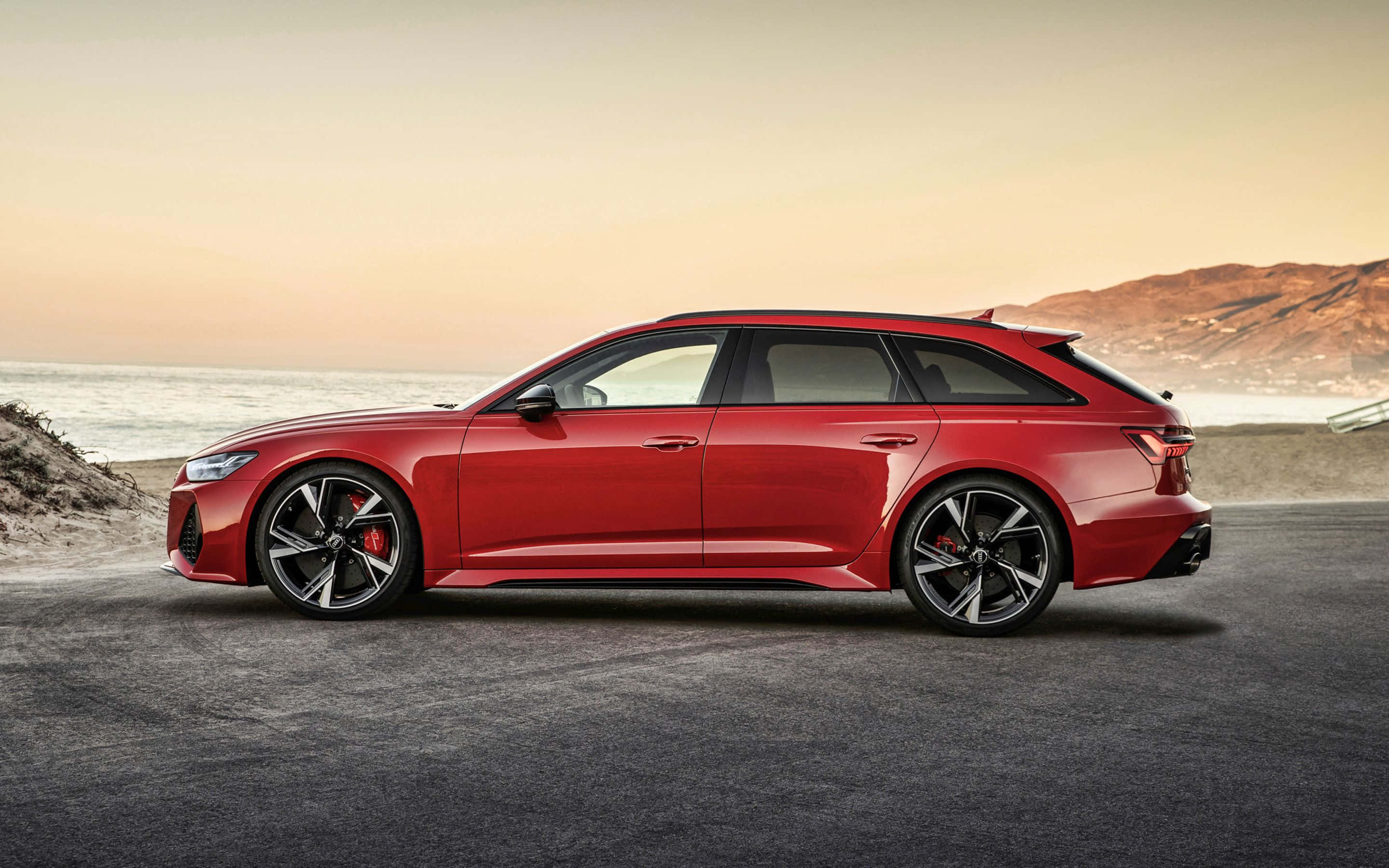 Audi Rs6 桌布 壁紙 — 一輛時尚的紅色Audi RS6 Avant旅行車停泊在日落時分的海岸公路上，背景是海洋和山丘。