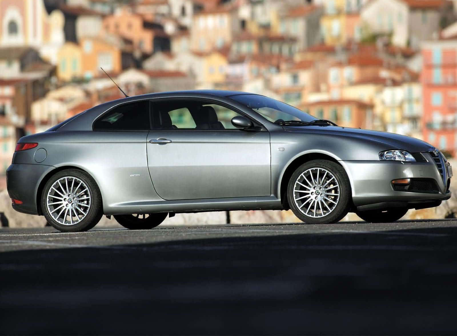 Alfa Romeo Gt 桌布 壁紙 — 一架時尚的銀色愛快羅密歐雙門跑車停泊在道路上，背景是色彩繽紛的山城，呈現模糊效果。