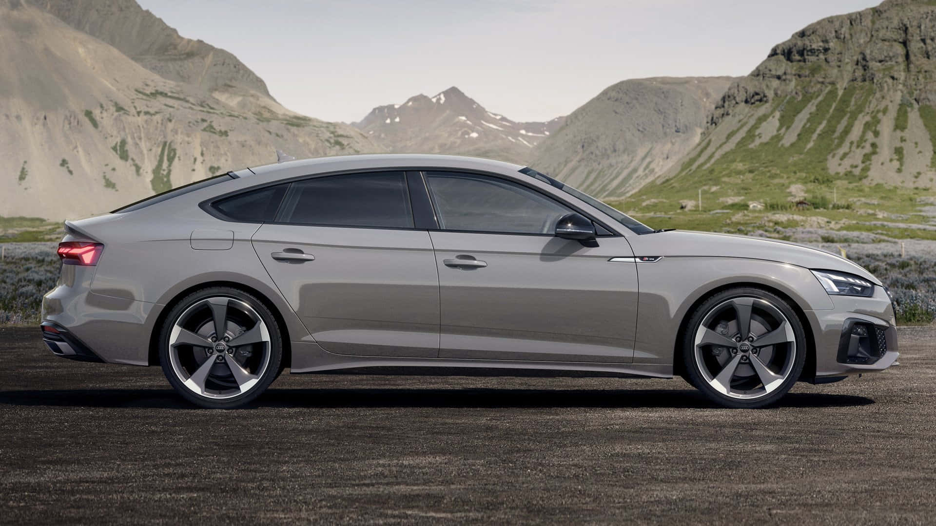 Audi A5 桌布 壁紙 — 一輛時尚的銀色奧迪轎車停在碎石上，背景是崎嶇的山脈和稀疏的綠植，天空明亮。