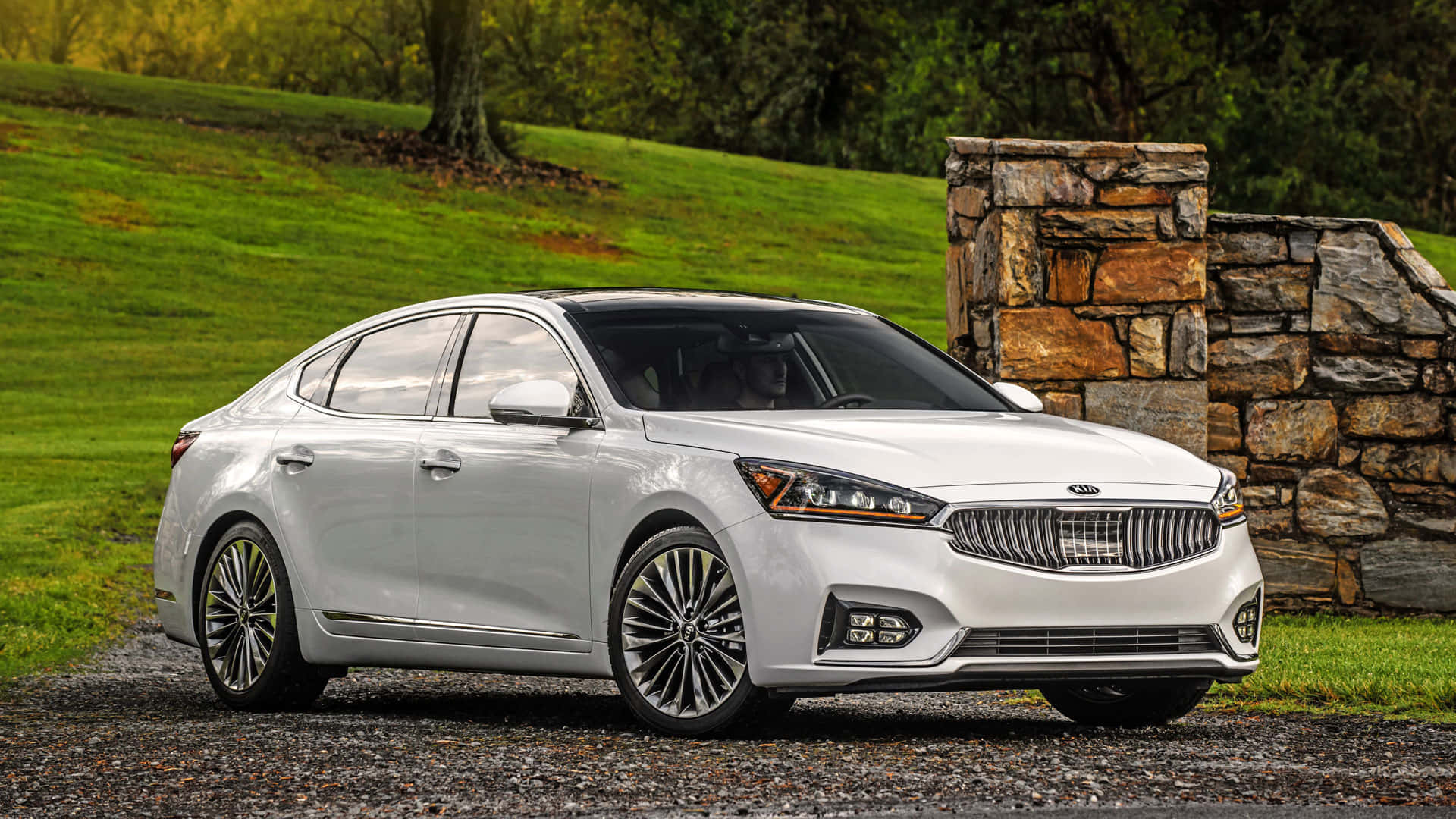 Kia Cadenza 桌布 壁紙 — 一輛時尚的銀色Kia轎車停放在碎石路上，旁邊是石牆，背景有茂密的綠草和樹木。