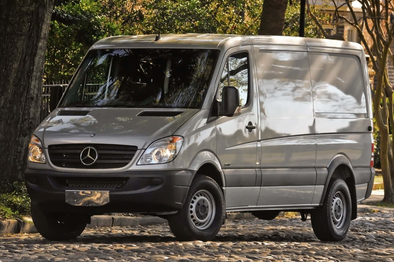 Mercedes Benz Sprinter 桌布 壁紙 — 一輛時尚的銀色梅賽德斯-奔馳 Sprinter 客貨車停放在陽光普照、樹木成蔭的鵝卵石街道上。