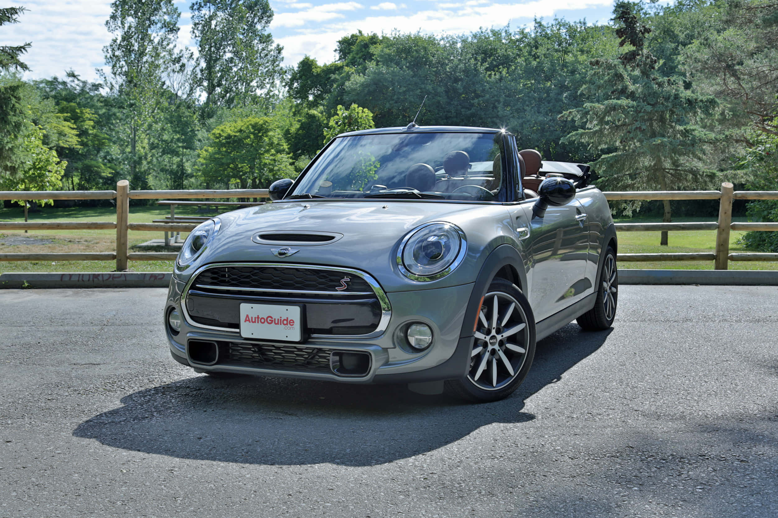 Mini Cooper S Convertible 桌布 壁紙 — 一輛時尚的銀色MINI Cooper敞篷車停在柏油路上，背景有茂密的綠樹和木柵欄，在明亮的日光下。