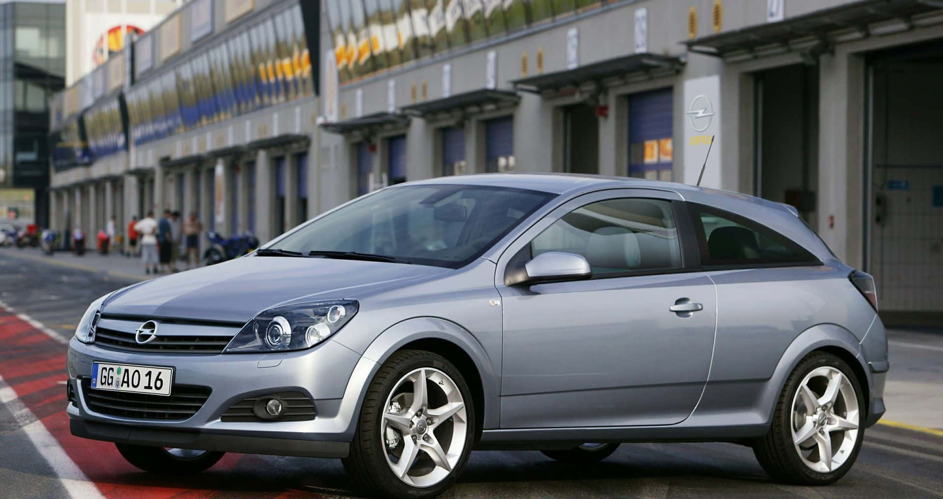 Opel Astra 桌布 壁紙 — 一輛時尚的銀色歐寶雅特雙門轎跑車停泊在都市街道上，背景有現代建築物和行人。