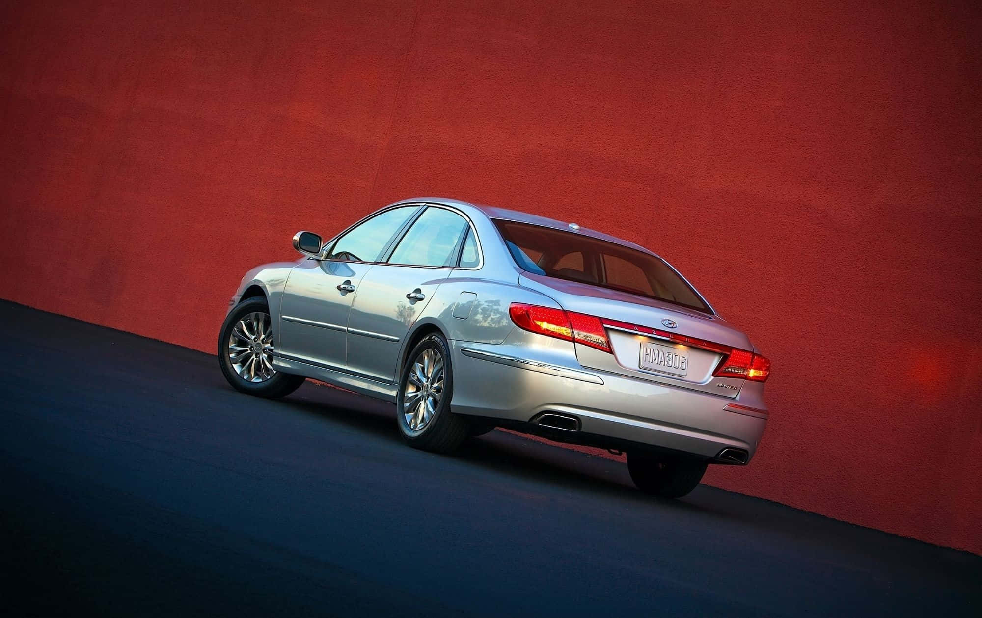 Hyundai Azera 桌布 壁紙 — 一輛時尚的銀色轎車在攝影棚燈光下傾斜靠在有紋理的紅色牆壁上。