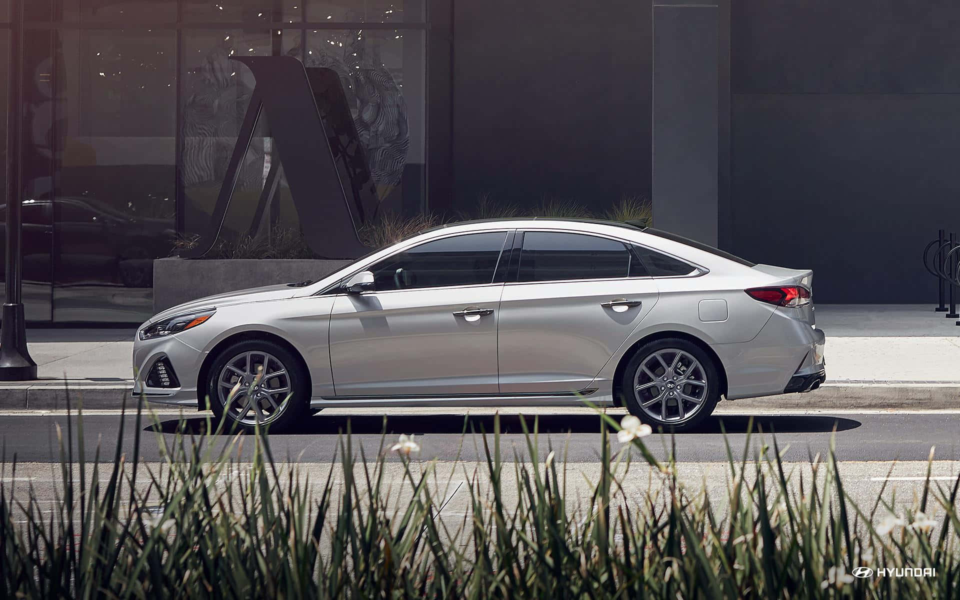 Hyundai Sonata 桌布 壁紙 — 一輛時尚的銀色轎車停泊在現代玻璃建築和綠化前景的城市街道上。