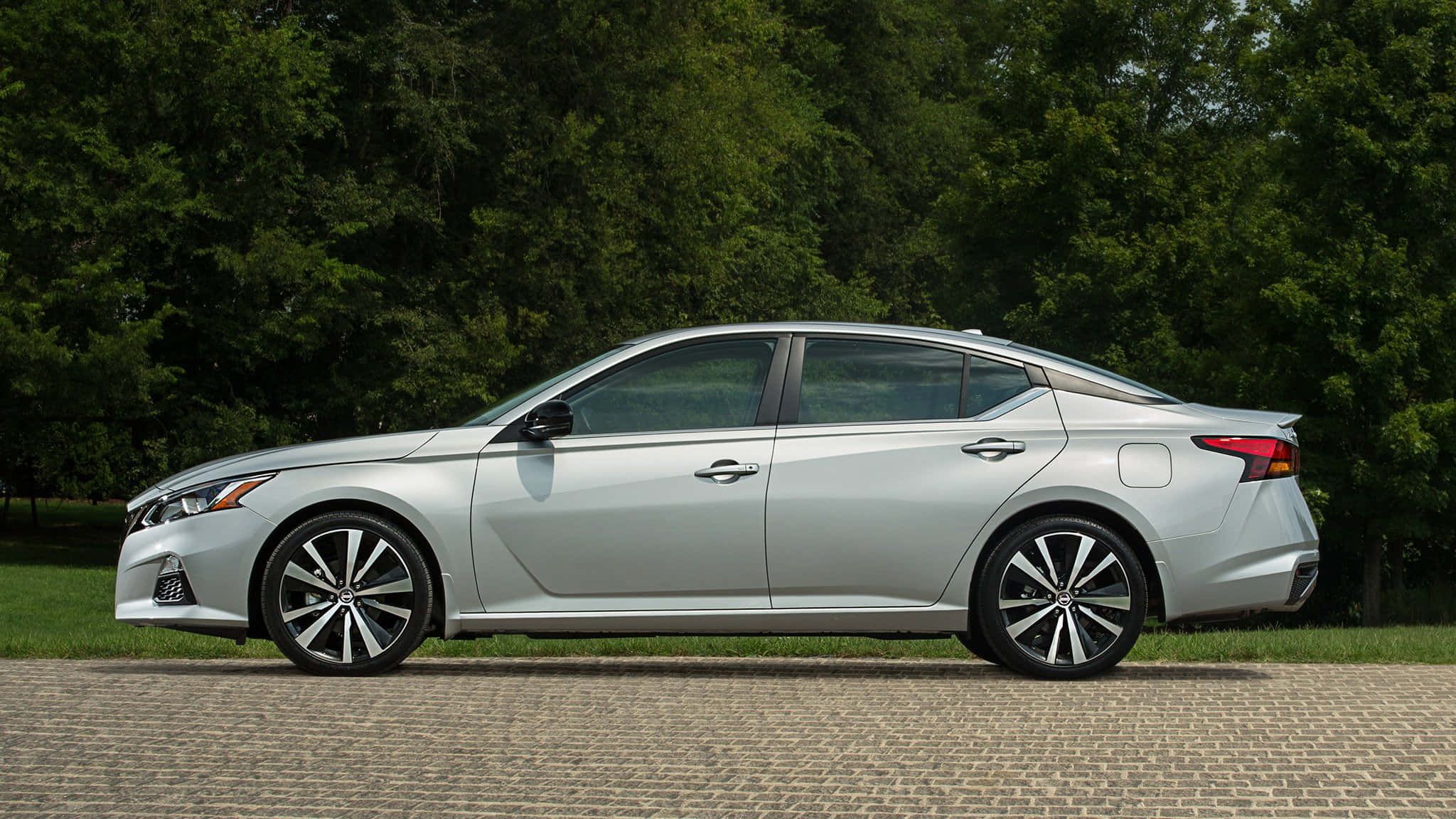 Nissan Altima 桌布 壁紙 — 一輛時尚的銀色轎車停泊在鵝卵石小徑上，背景是茂密的綠樹。