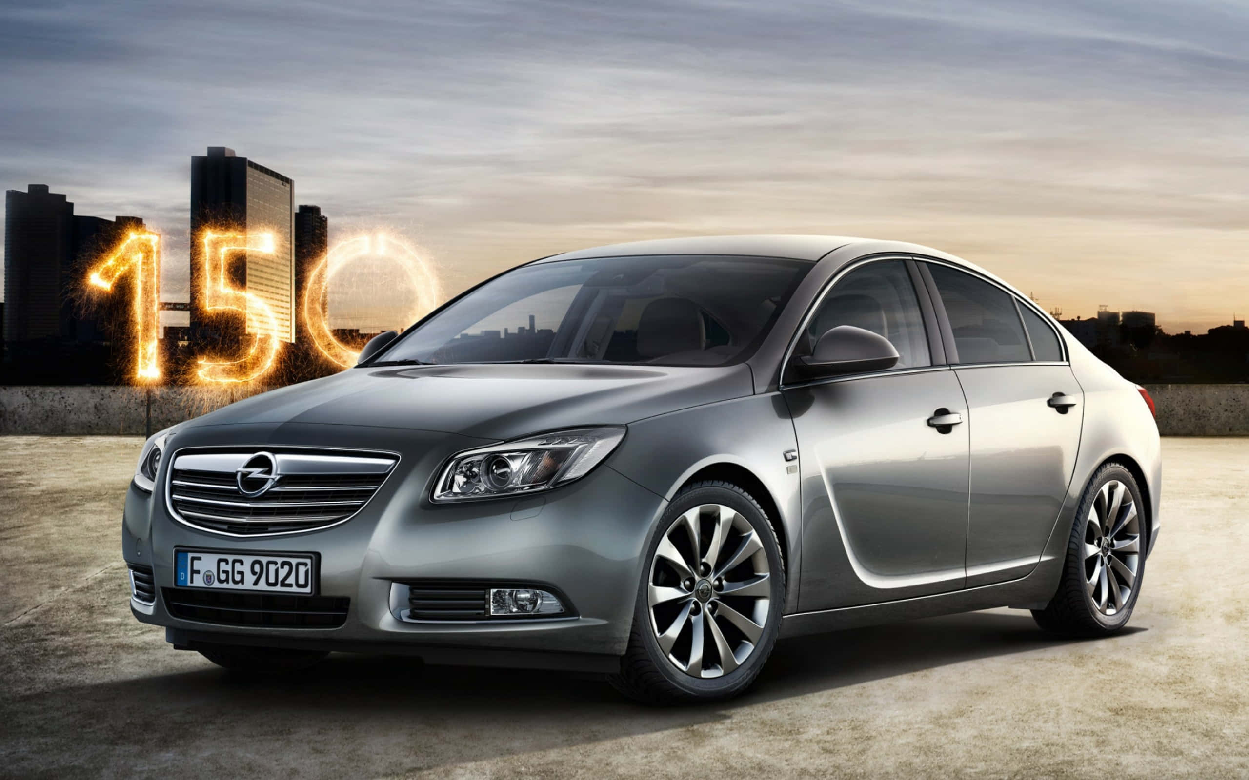 Opel Insignia 桌布 壁紙 — 一輛時尚的銀色轎車停在日落時分的屋頂上，背後是發光的「150」煙火，襯托著城市天際線。