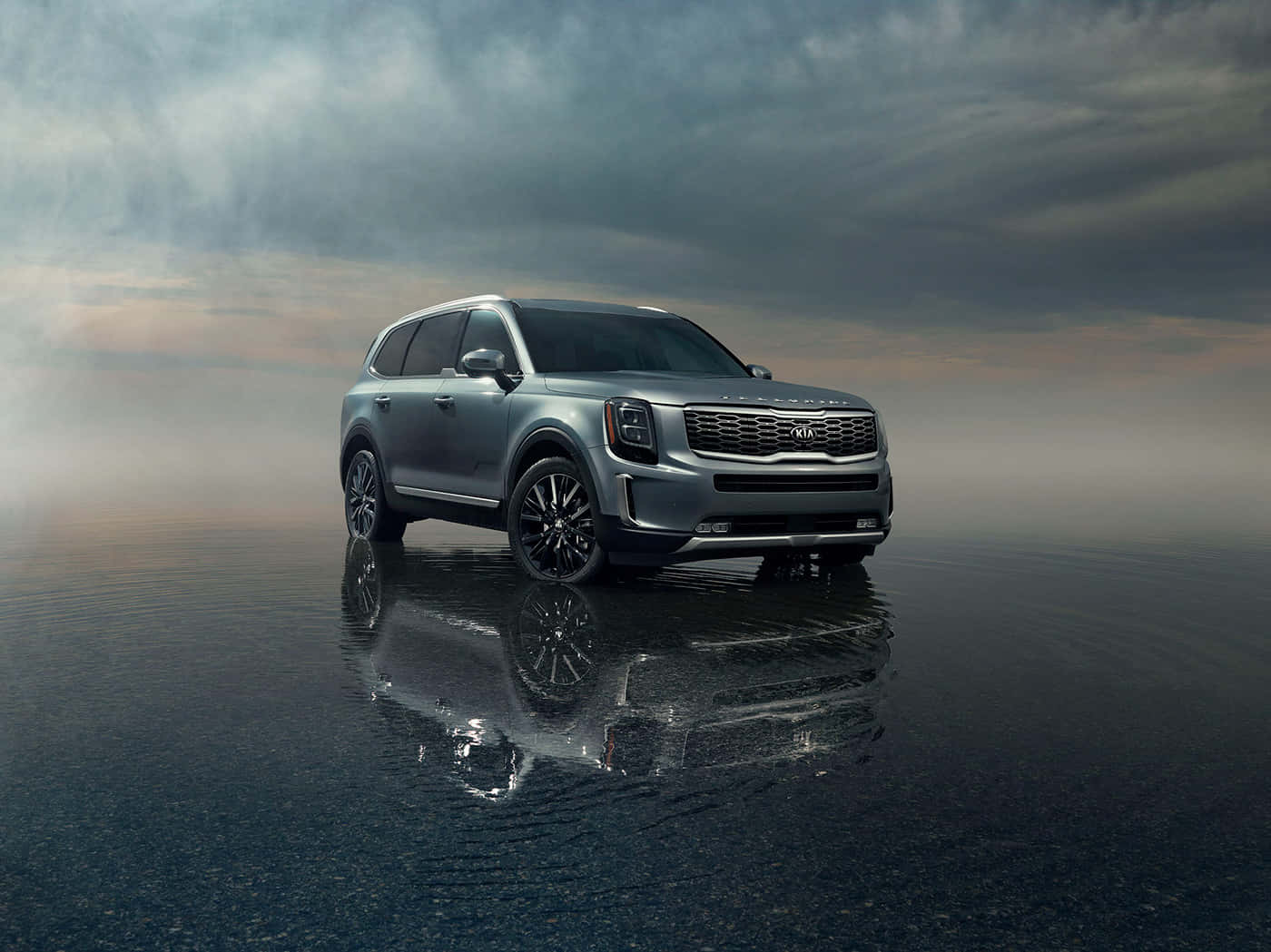 Kia Telluride 桌布 壁紙 — 一輛時尚的銀色SUV佇立在反光的濕滑路面上，背景是充滿戲劇性雲層的暮色天空。