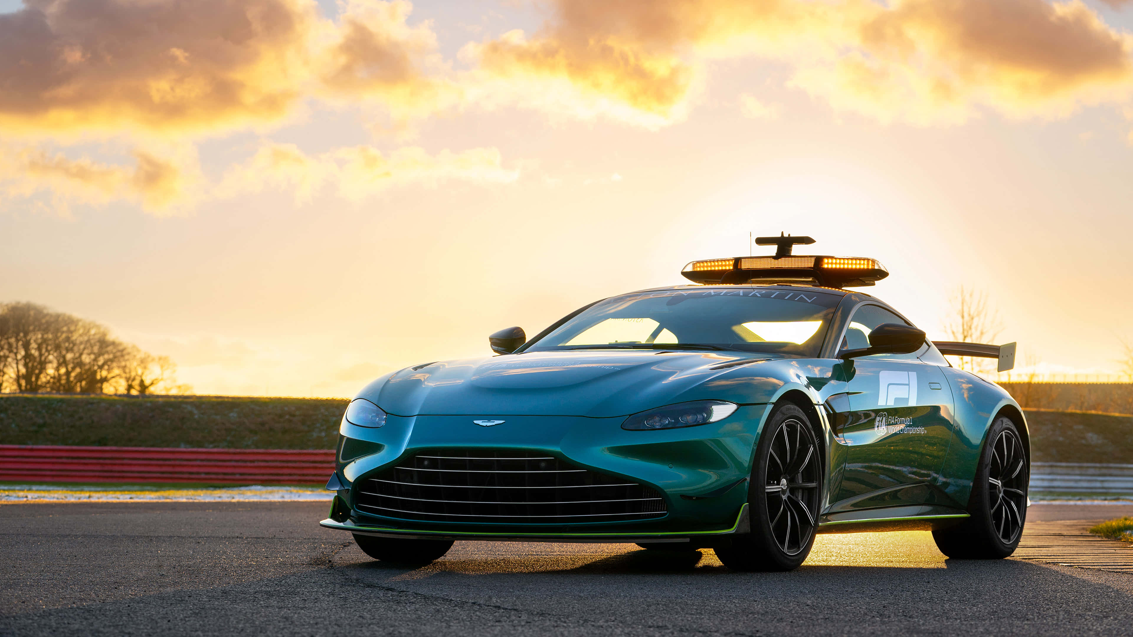 Aston Martin Vantage 桌布 壁紙 — 一架時尚的藍綠色 Aston Martin 賽車，車頂裝有燈條，在金色夕陽下停在賽道上。
