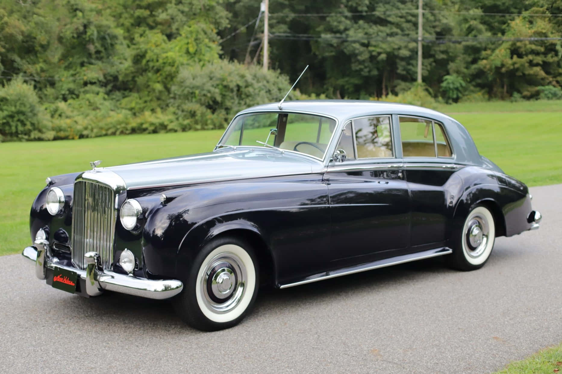 Bentley S2 桌布 壁紙 — 一輛時尚的復古賓利，採用黑色和銀色雙色調飾面，在日光下於一條兩旁長滿翠綠草地和樹木的柏油路上閃閃發光。