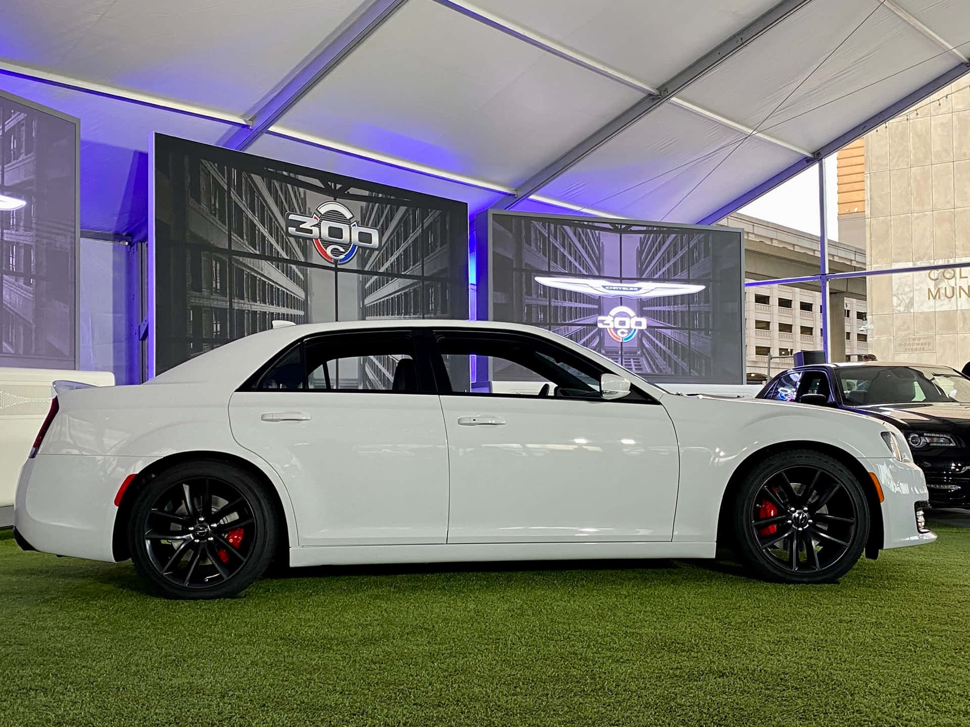 Chrysler 300 桌布 壁紙 — 一輛時尚的白色Chrysler 300轎車，配備黑色輪圈和紅色制動卡鉗，在帶有發光藍色燈光和品牌屏幕的帳篷下的綠色草坪上展示。