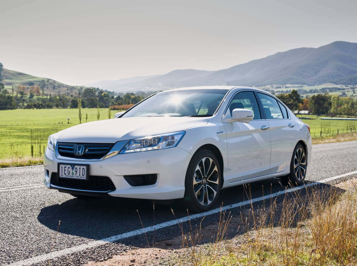 Honda Accord 桌布 壁紙 — 一輛時尚的白色本田雅閣轎車停泊在鄉村道路上，背景是起伏的綠色山丘和遠處的山脈，陽光明媚。