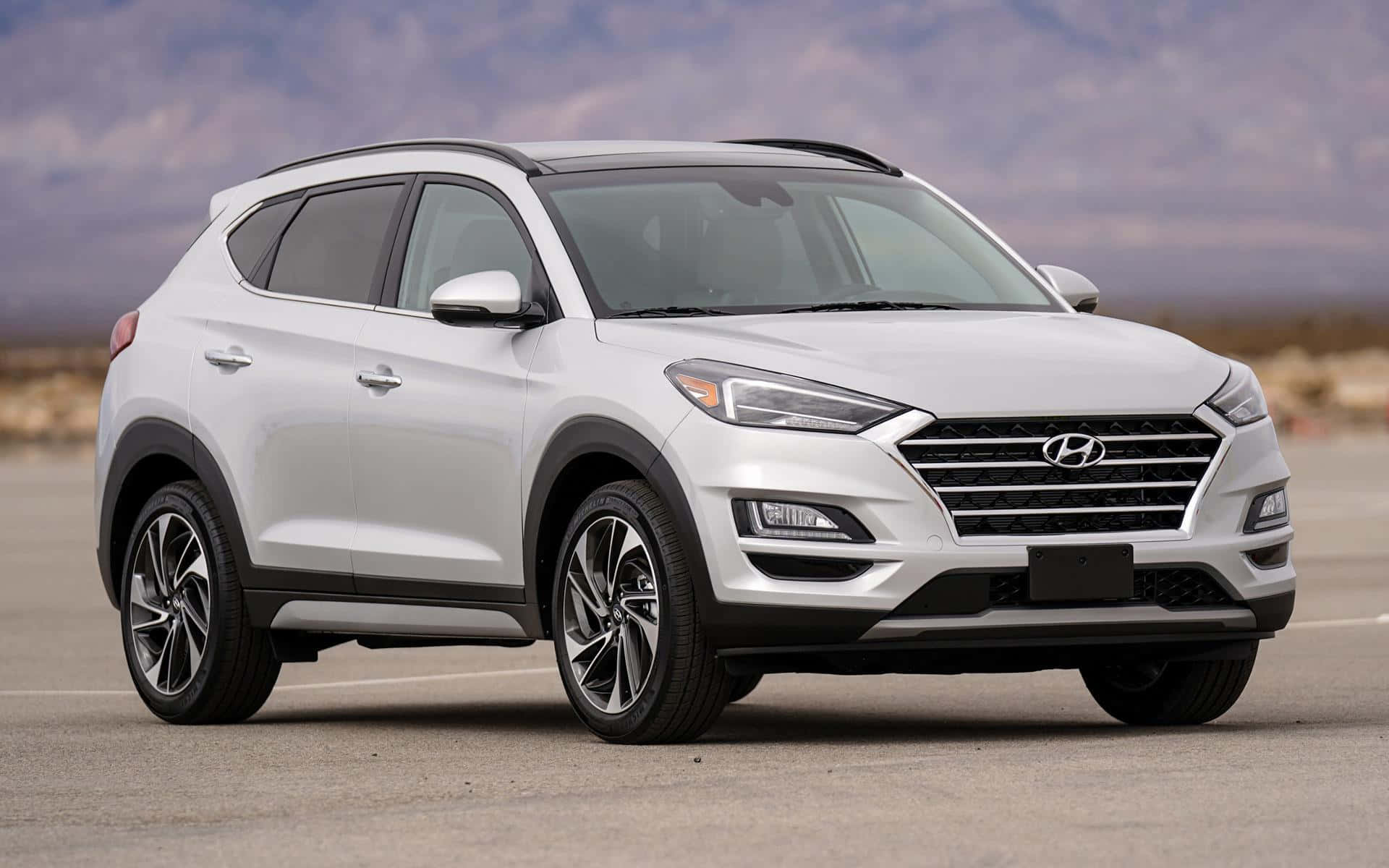 Hyundai Tucson 桌布 壁紙 — 一輛時尚的白色現代 Tucson SUV 停放在鋪砌的停車場上，背景是朦朧的山巒，天空多雲。