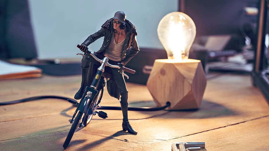 Aokiji 桌布 壁紙 — 一個風格化的動作人偶騎著腳踏車，坐在木地板上一個發光的燈泡燈旁邊。