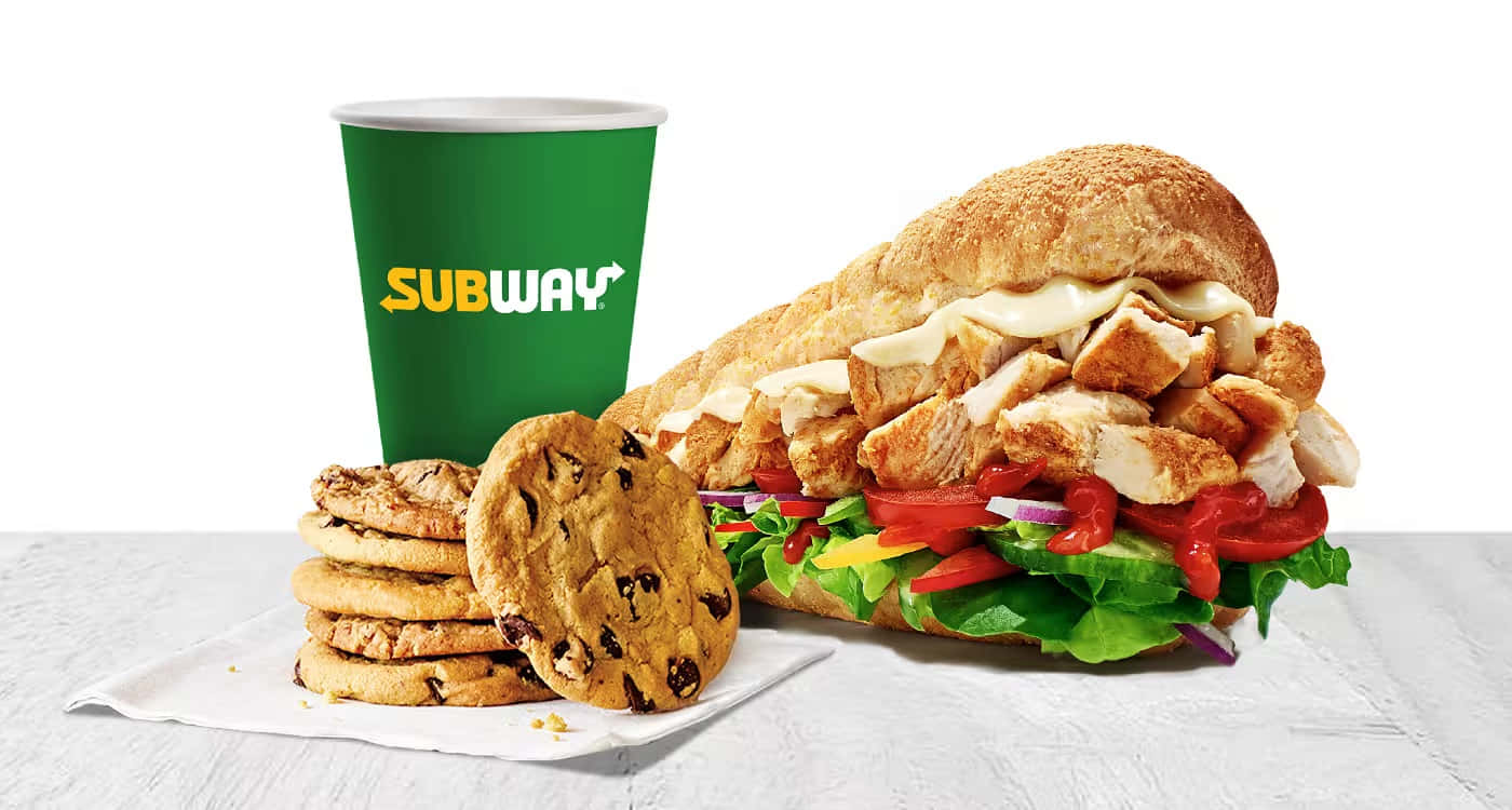 Subway 桌布 壁紙 — Subway 雞肉三明治搭配新鮮蔬菜，旁邊放著一個綠色飲料杯，還有一疊巧克力餅乾，放在白色紋理表面上。