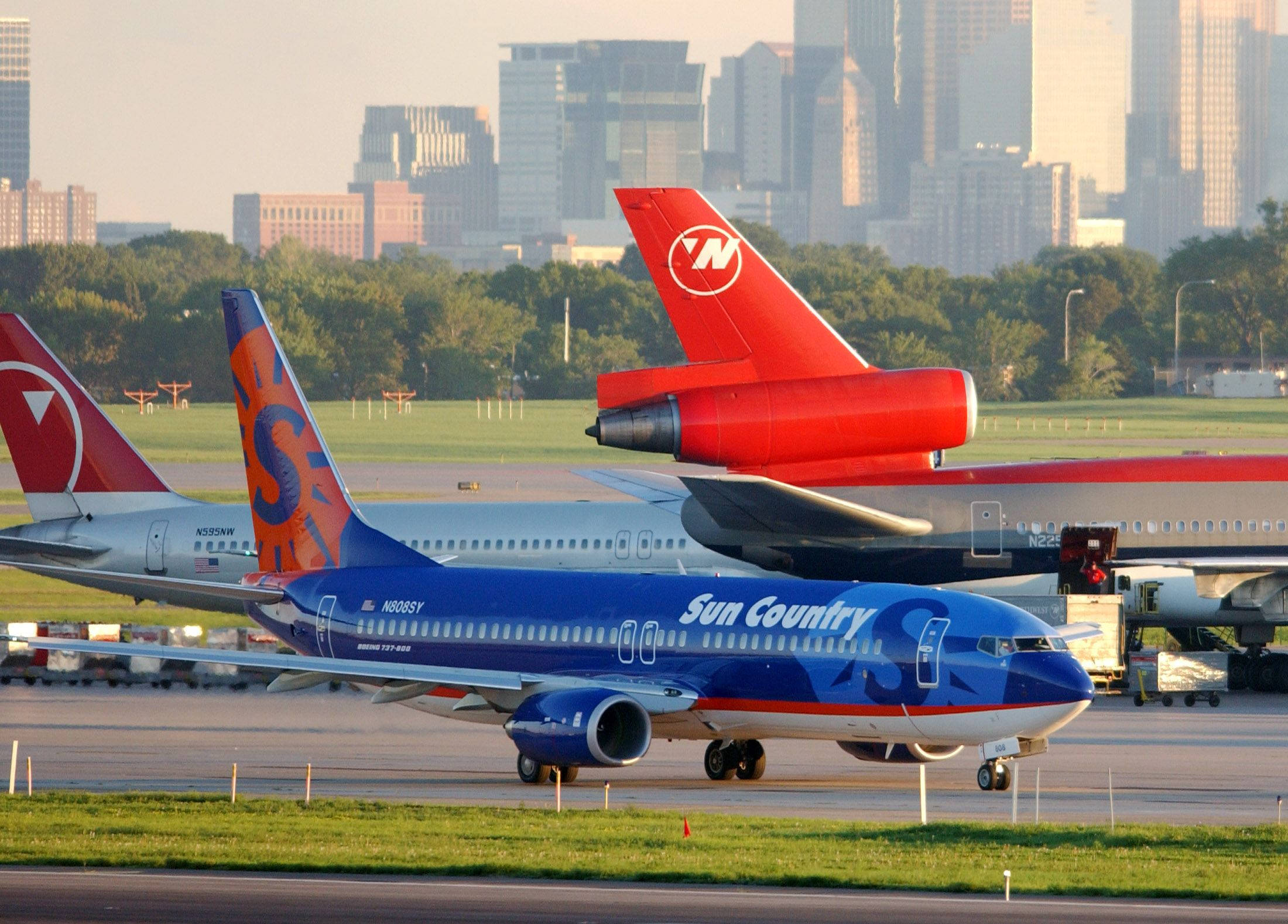 Airport 桌布 壁紙 — Sun Country Airlines 飛機在黃金時段於跑道上滑行，背景有城市天際線和其他噴射機。