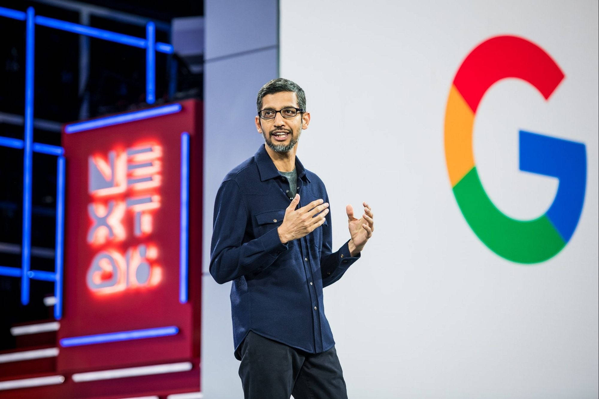 google 桌布 壁紙 — Sundar Pichai 在Google活動中發言時做手勢，身後有一個大型彩色G標誌和發光的「NEXT」標牌。