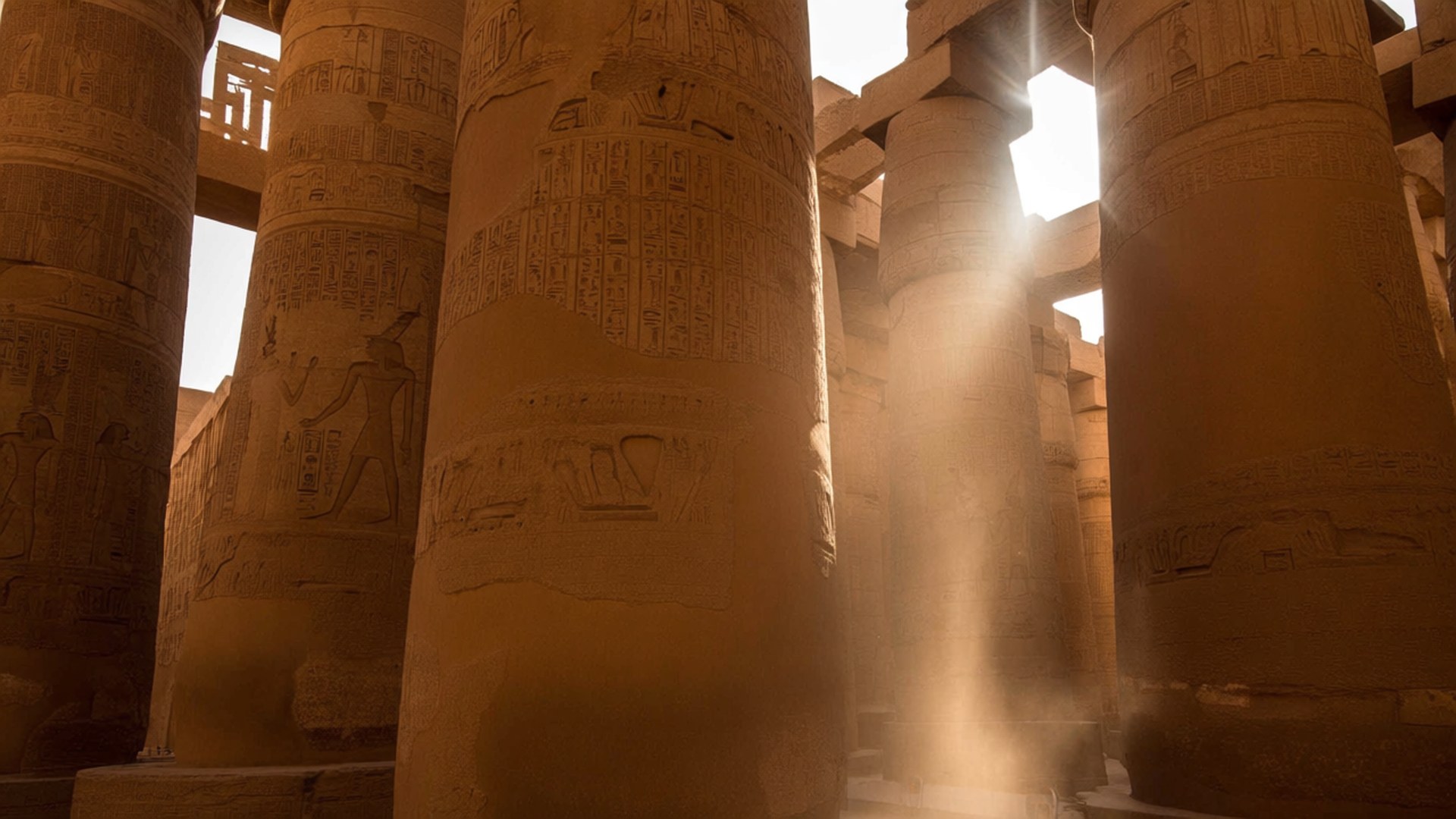 Karnak Sunbeam 壁紙 — 陽光穿過刻有象形文字和人物的古埃及神廟柱子，在溫暖的琥珀色光線中揚起金色的塵埃。