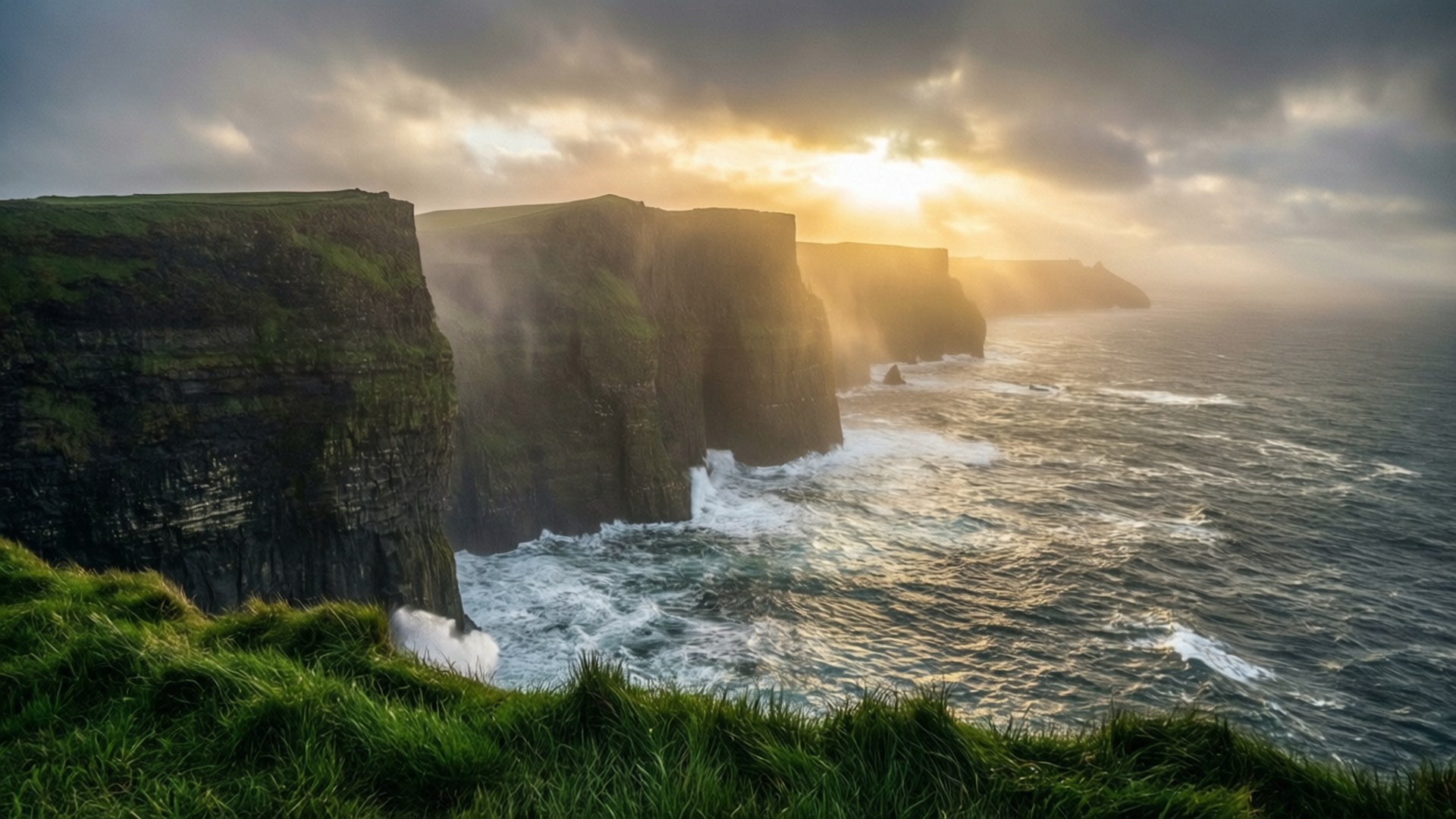 Cliffs of Moher Sunrise 壁紙 — 陽光穿過暴風雲，照亮了戲劇性的海崖，海浪在茂密的綠草下拍打。