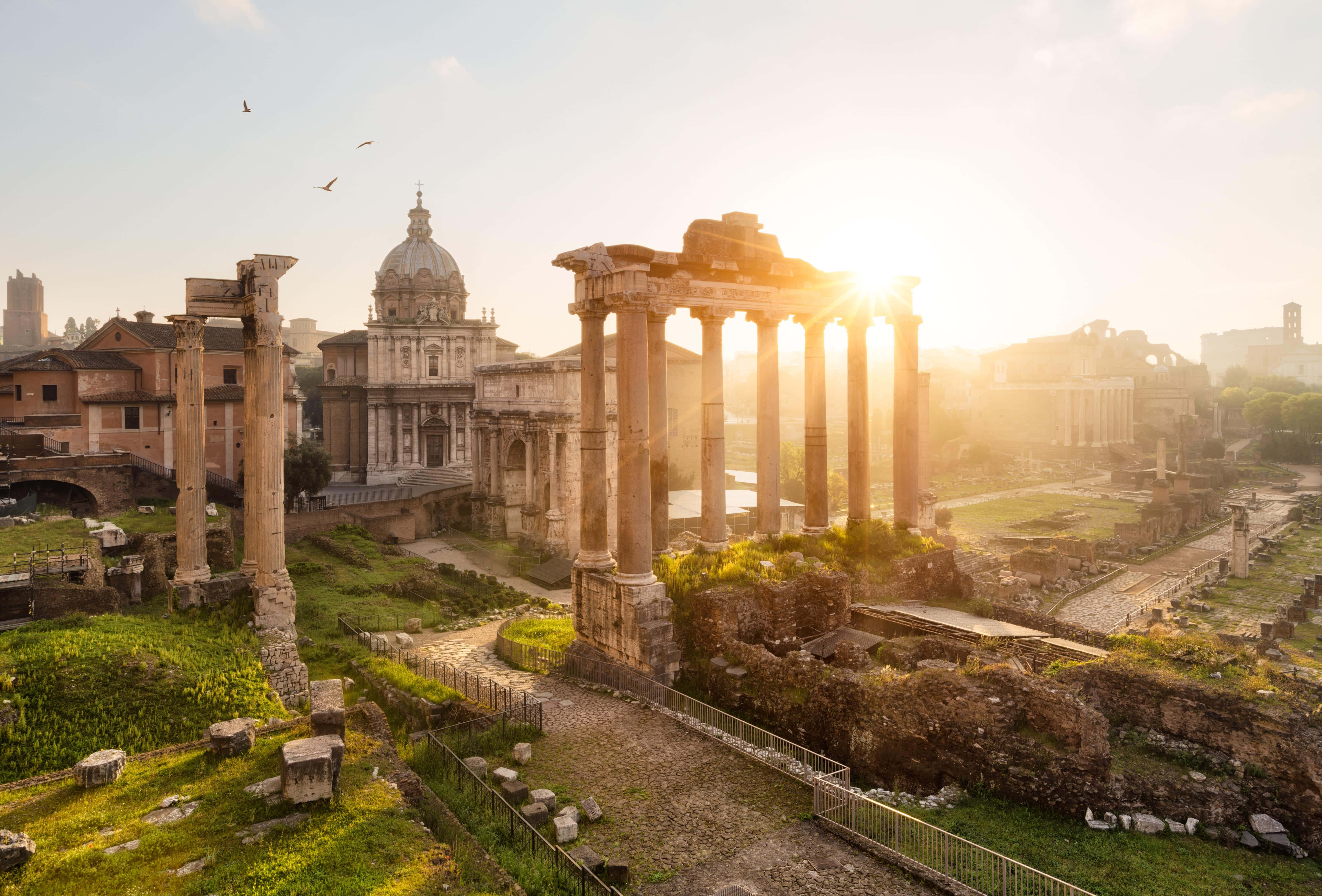 Roman 桌布 壁紙 — 陽光穿過古羅馬柱，在長滿草的廢墟和遠處的圓頂教堂上投下長長的影子。