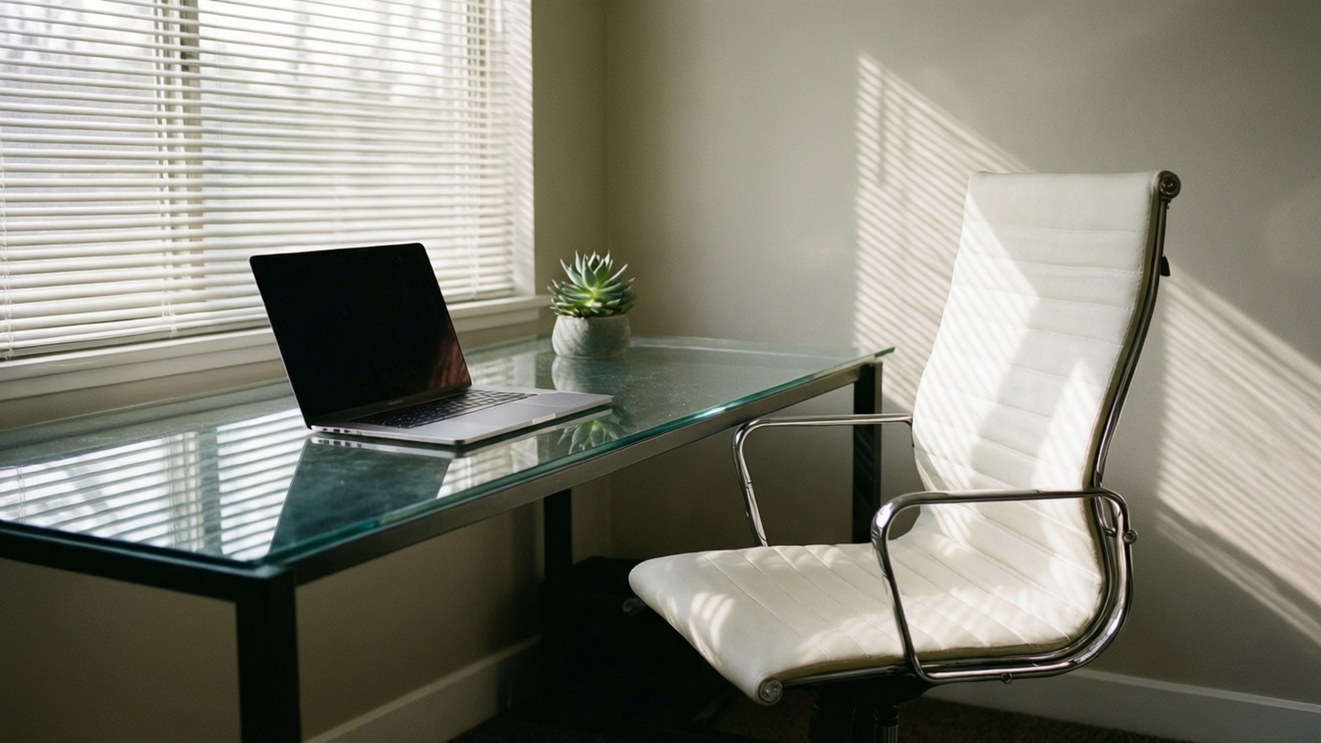 Modern Home Office 壁紙 — 陽光穿過百葉窗，在玻璃桌上投射出條紋陰影，桌上有一台筆記型電腦和一棵多肉植物，旁邊是一張白色皮革椅子。