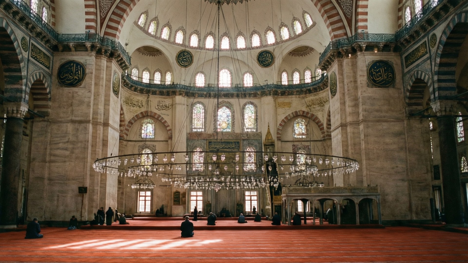 Süleymaniye Interior 壁紙 — 陽光透過彩色玻璃窗照射進來，照亮了宏偉圓頂清真寺內紅地毯上的禮拜者，清真寺內有複雜的瓷磚工藝和懸掛的吊燈。