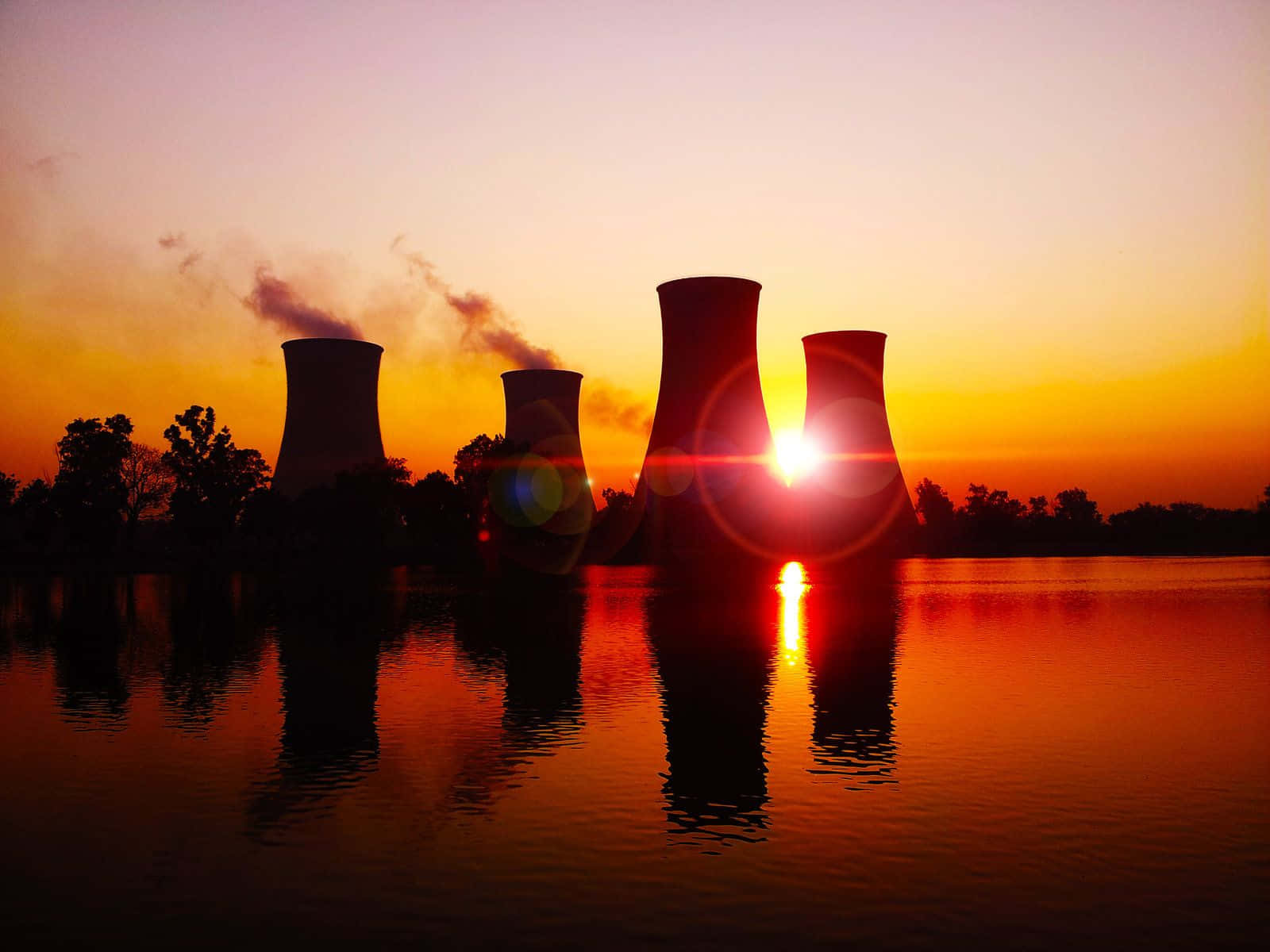  壁紙 — 夕陽在剪影般的核電廠冷卻塔後發光，倒映在平靜的水面上，煙霧升起。