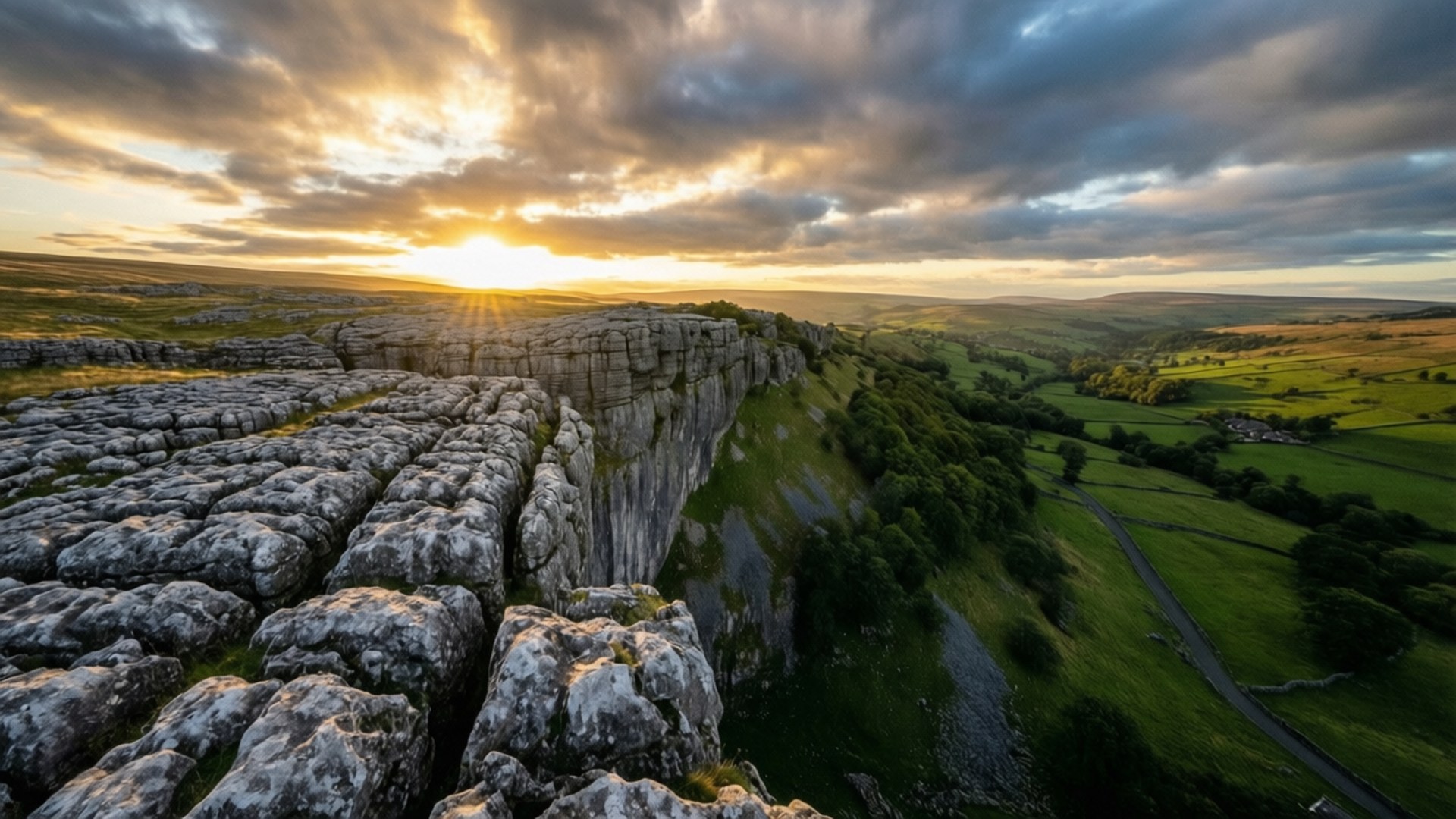 Sunrise Over Malham Cove 壁紙 — 夕陽照亮了戲劇性的石灰岩懸崖邊緣，俯瞰著鬱鬱蔥蔥的綠色山谷和蜿蜒的道路。