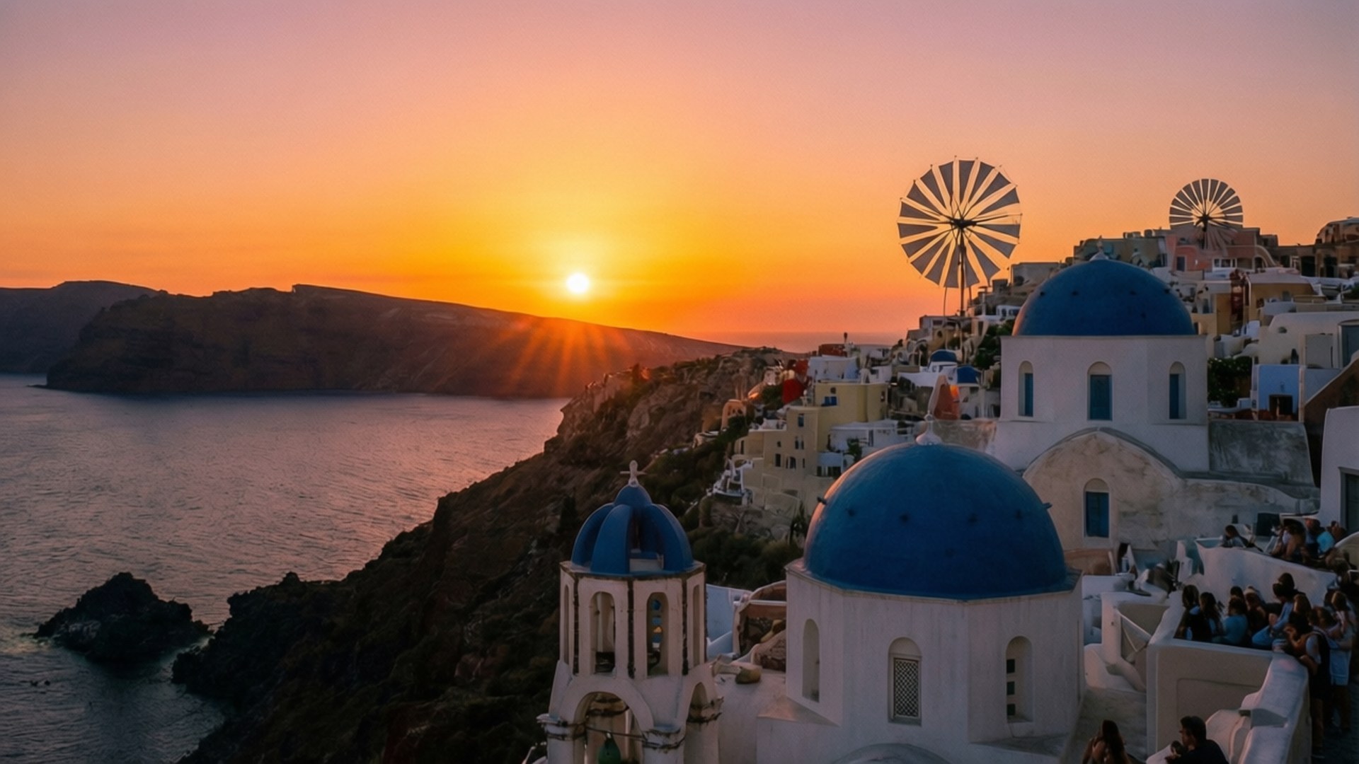 Oia Sunset Romance 壁紙 — 聖托里尼的日落，藍色圓頂教堂、白色建築和風車俯瞰愛琴海。
