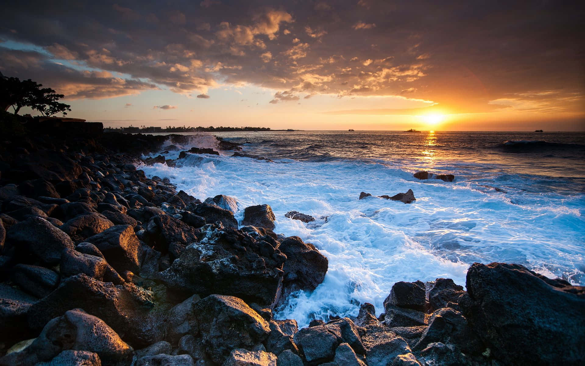 Coastal Sunset 桌布 壁紙 — 夕陽將天空染成火熱的橙色，海浪拍打著崎嶇海岸線上的深色火山岩。