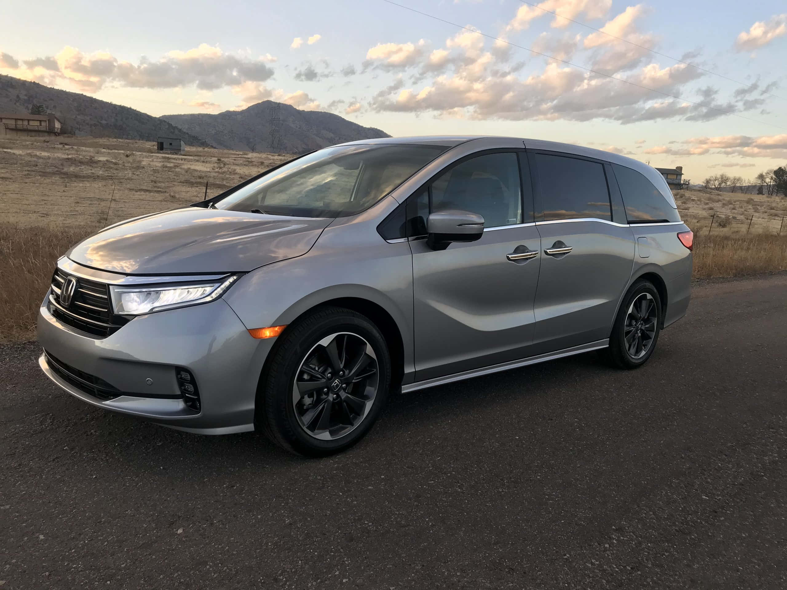 Honda Odyssey 桌布 壁紙 — 一輛時尚的銀色小型貨車停泊在鄉村道路上，背景是山脈和金色的雲彩，正值日落時分。