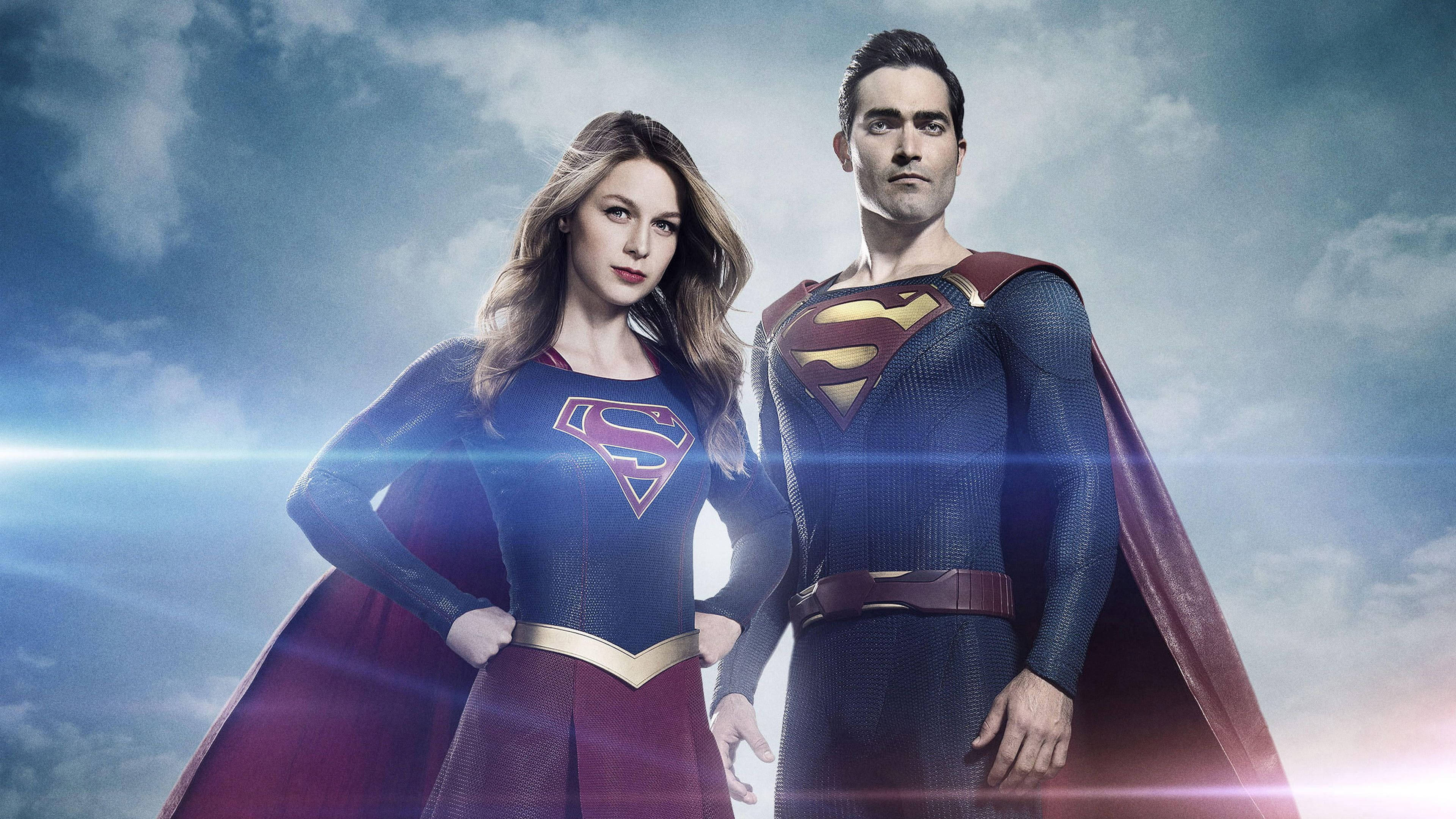 Superwoman 桌布 壁紙 — Supergirl 和 Superman 自信地站在戲劇性的多雲天空下，明亮的鏡頭光暈照亮了他們標誌性的藍色和紅色服裝。