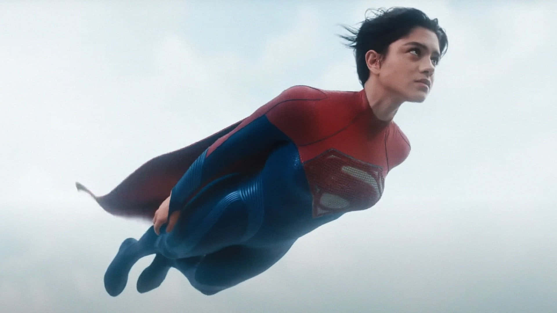  壁紙 — 一位身穿紅藍戰衣的超級英雄，披著飄逸的斗篷，帶著堅定的表情，在陰雲密佈的天空中翱翔。