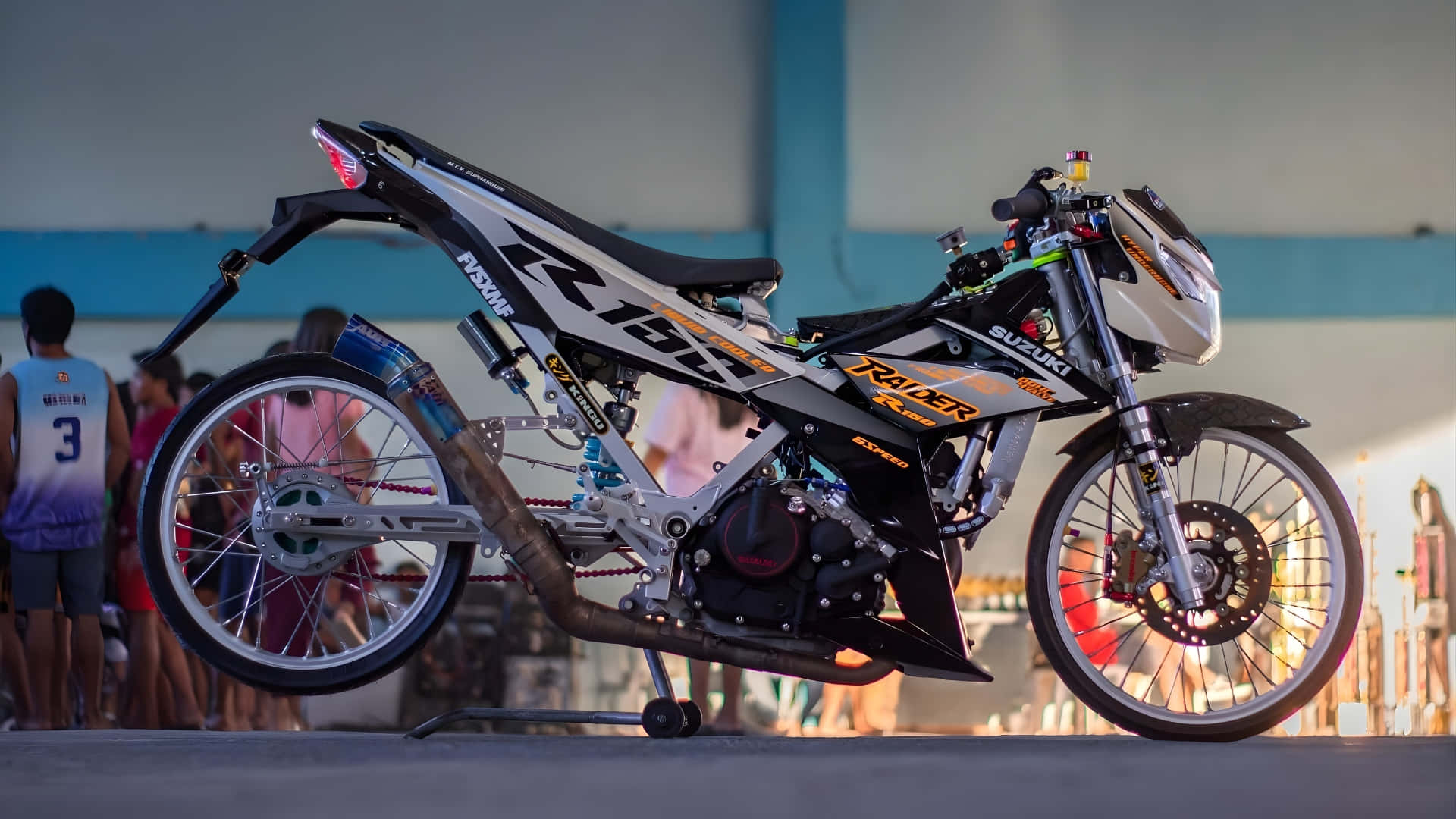  壁紙 — 一架時尚的黑白 Suzuki 越野機車帶有橙色點綴，在黃昏時立於支架上，背景有模糊的觀眾。