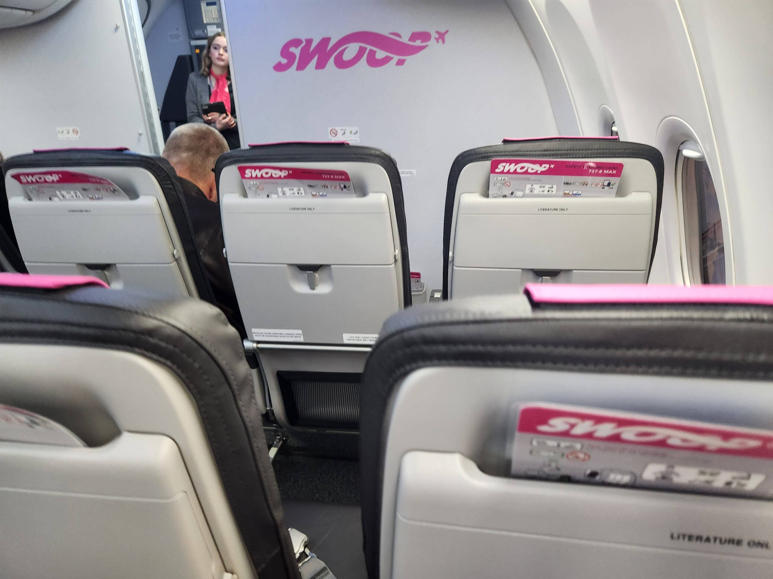 seat 桌布 壁紙 — Swoop 飛機艙內部，顯示一排排灰色和黑色座椅帶有粉紅色點綴，背景中有一名空服員。