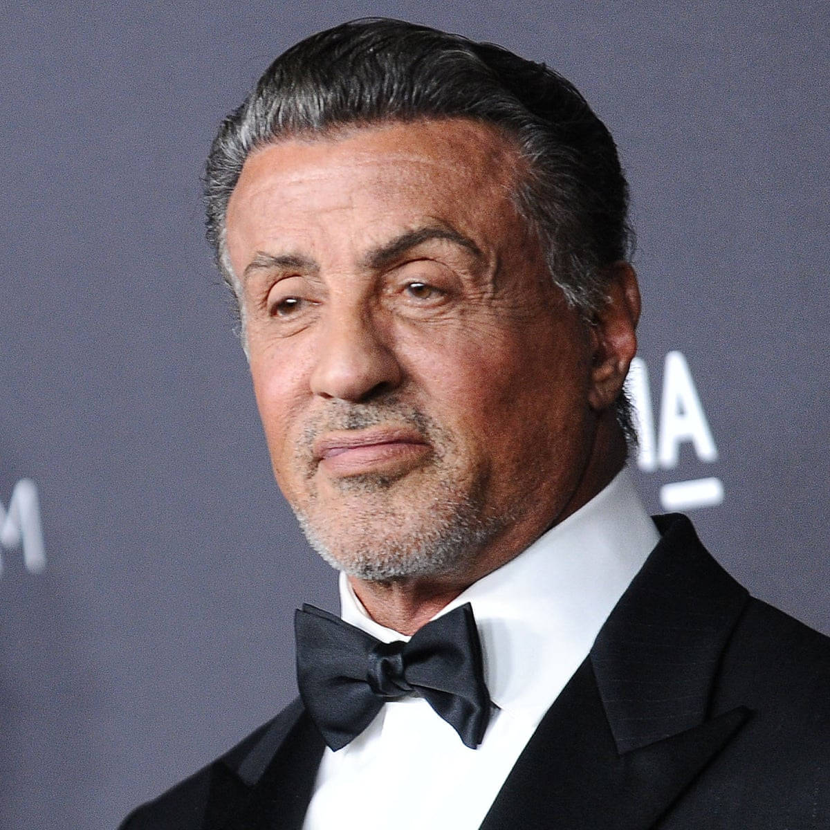 iPhone 桌布 壁紙 — Sylvester Stallone 身穿黑色修身燕尾服和領結，在灰色活動背景前擺出嚴肅的表情。
