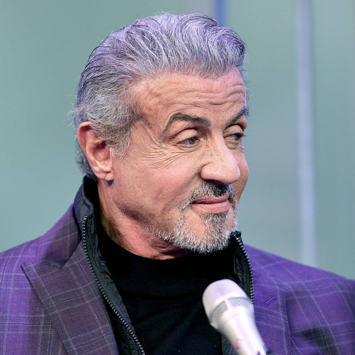 手機 桌布 壁紙 — Sylvester Stallone 灰白頭髮和鬍鬚，微笑著對著麥克風說話，身穿紫色格子外套。