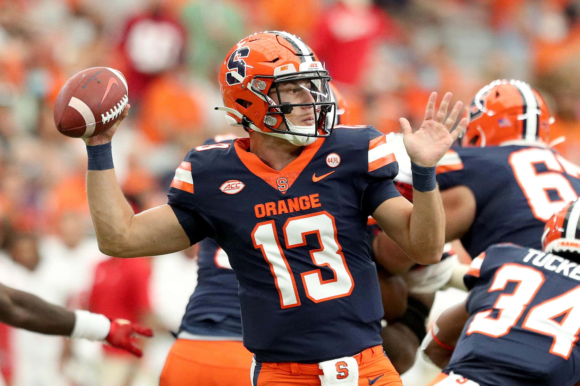  壁紙 — 一位身穿海軍藍和橙色球衣的 Syracuse Orange 美式足球四分衛正在日間比賽中準備傳球。