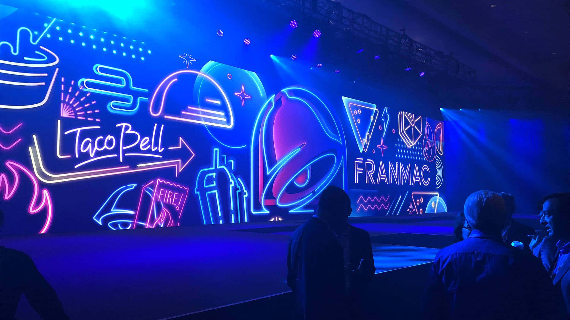 塔可鐘 桌布 壁紙 — Taco Bell 和 Franmao 的鮮豔霓虹燈牌在舞台上閃耀，剪影般的觀眾在戲劇性的藍色燈光下觀看。