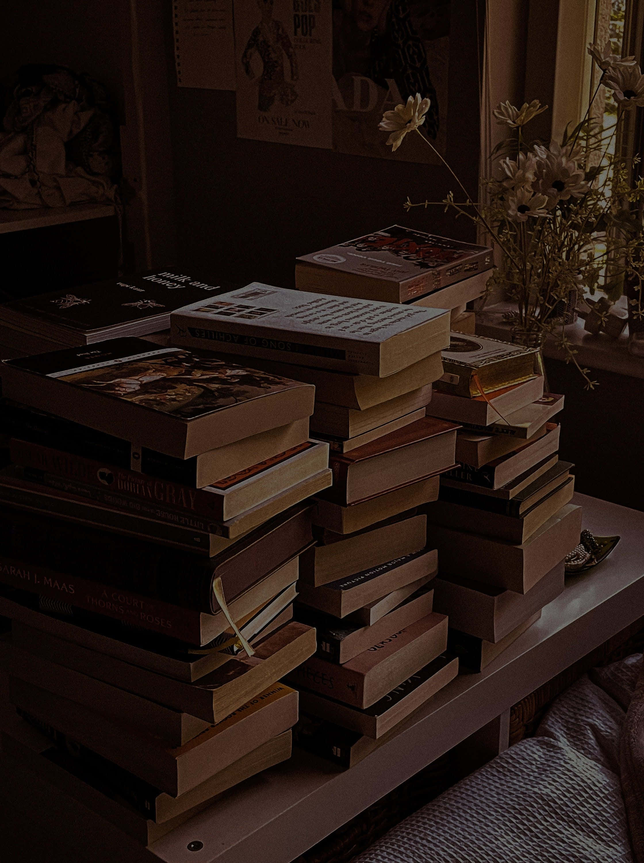 Dark Book Aesthetic 桌布 壁紙 — 高高的書堆放在木桌上，旁邊是窗戶，窗戶上有乾花和海報，房間光線昏暗。