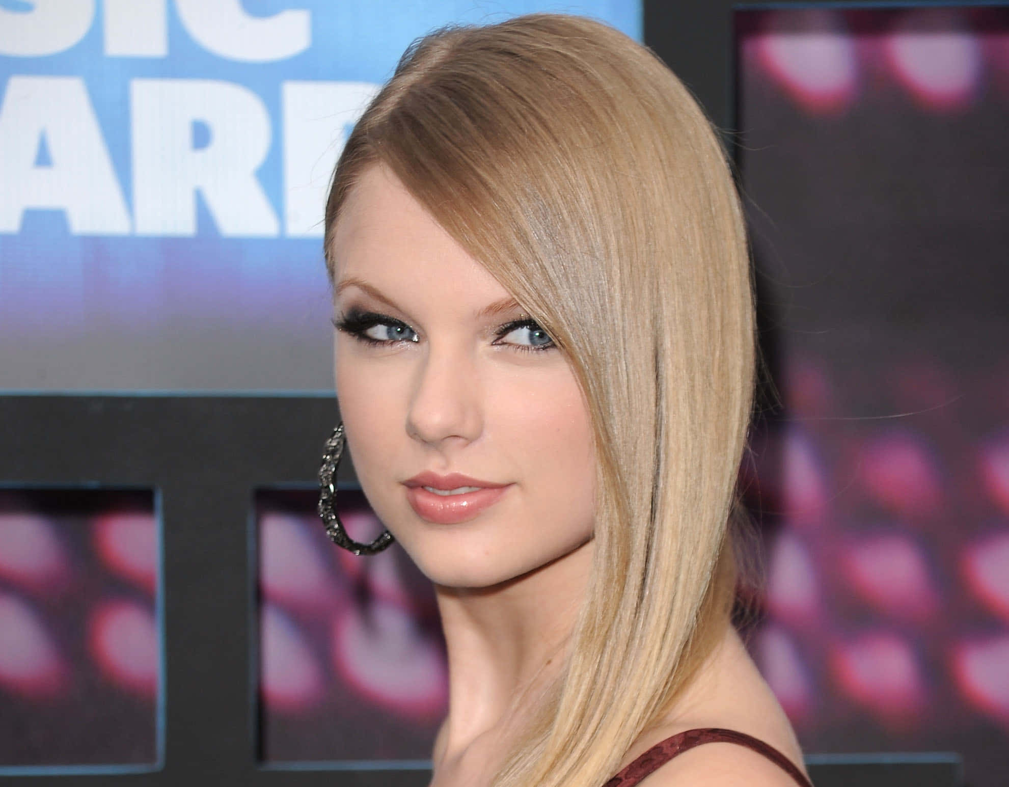 泰勒絲 桌布 壁紙 — Taylor Swift 以金髮和耳環造型在音樂頒獎活動中擺姿勢，身後有模糊的粉紅燈光。